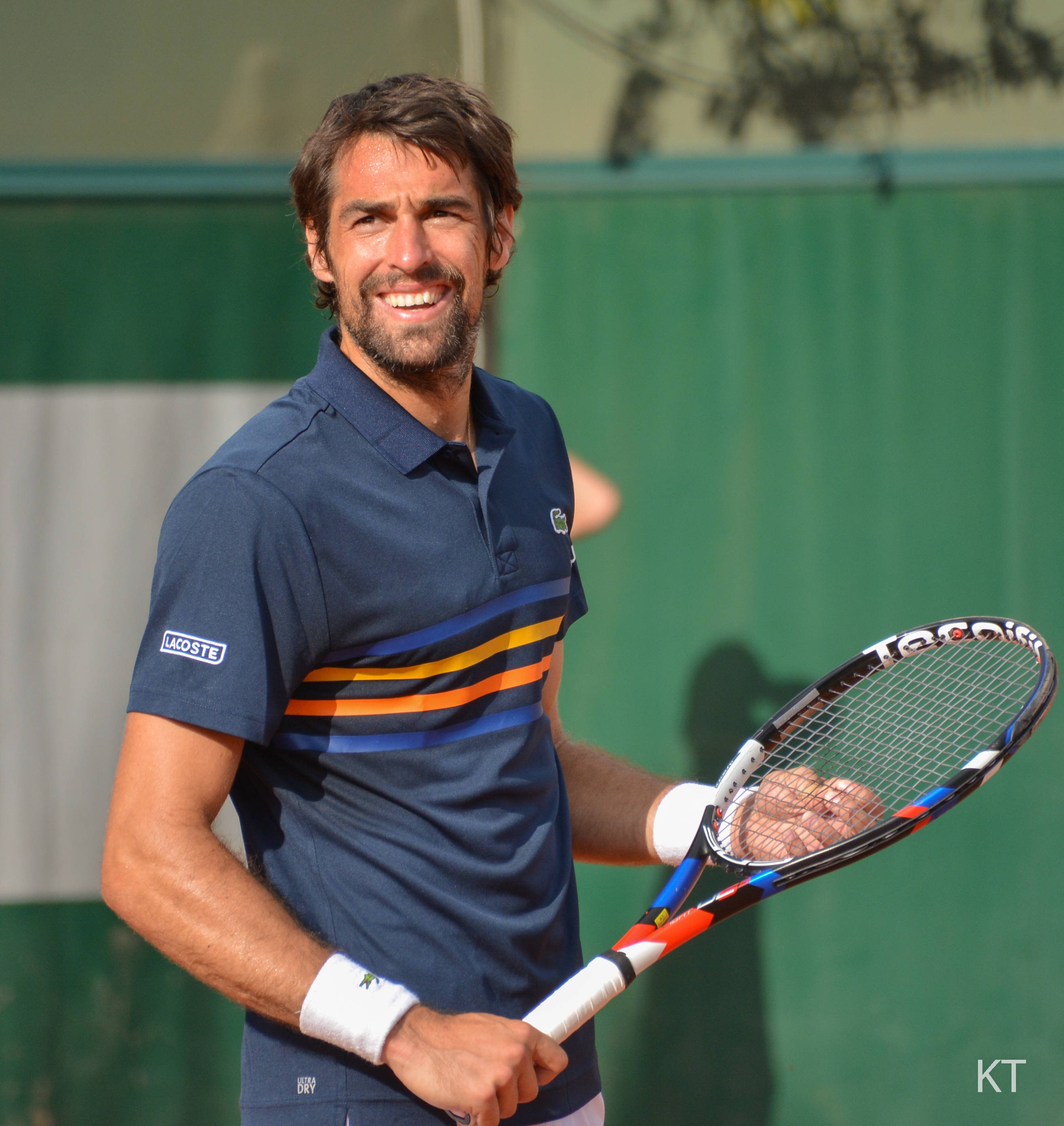 Tennis 桌布 壁紙 — 一位微笑的網球選手身穿藍色Lacoste Polo衫，帶有橙色和黃色條紋，在明亮的陽光下對著綠色圍欄握著球拍。