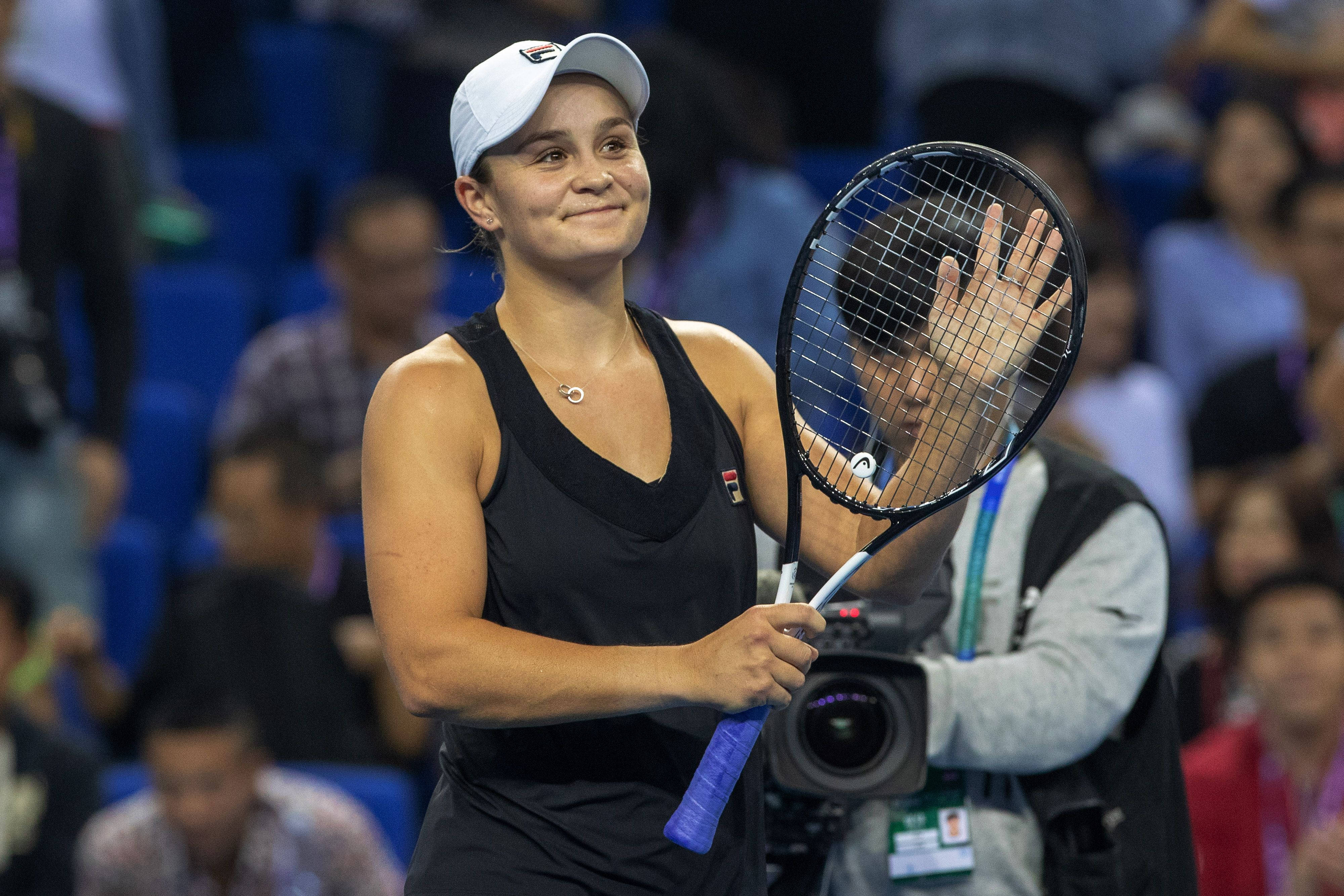 Ashleigh Barty 桌布 壁紙 — 一位微笑的網球選手身穿黑色服裝和白色帽子，在明亮的室內球場上拿著球拍向粉絲揮手。