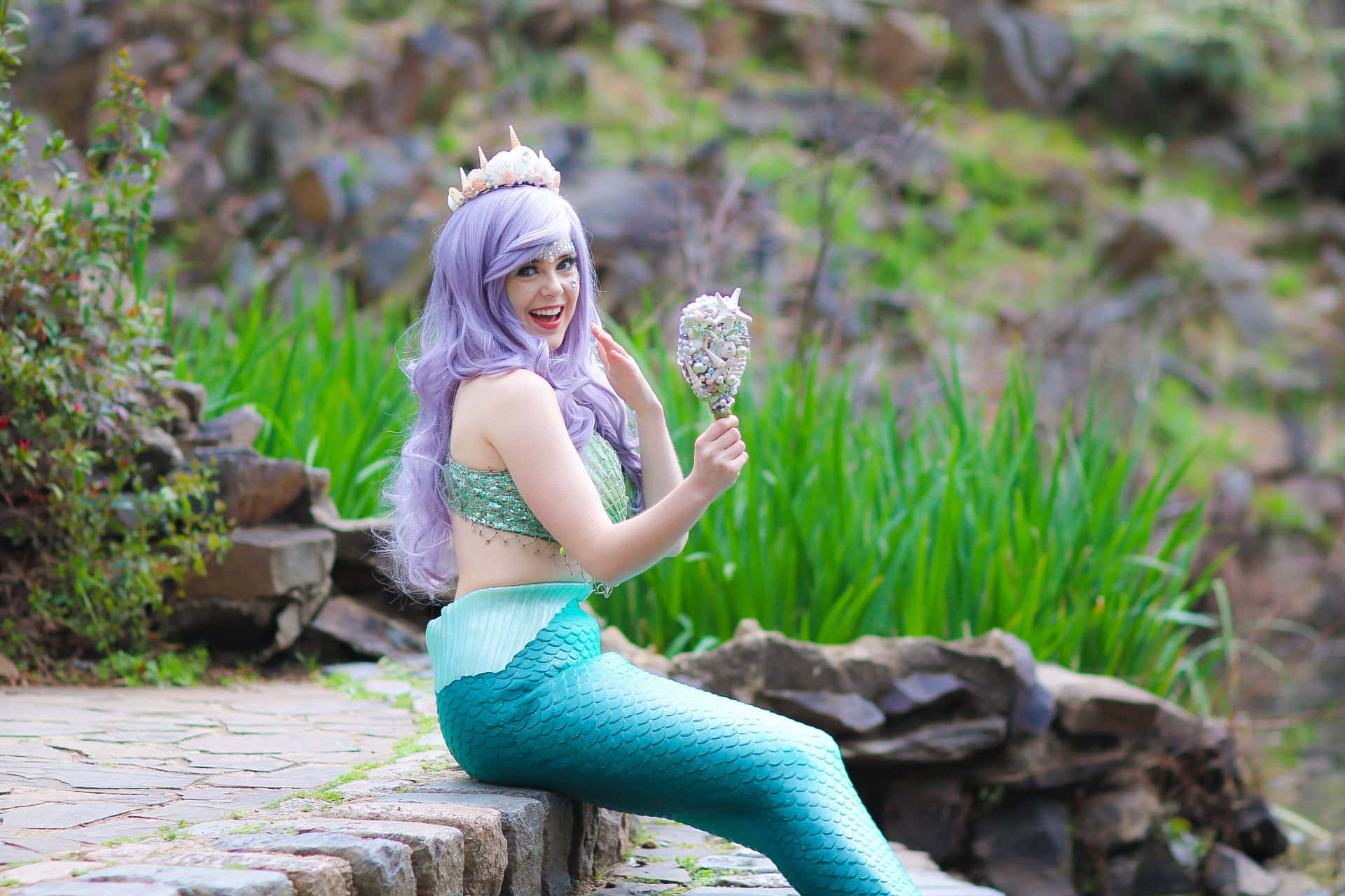 Real Mermaid 桌布 壁紙 — 一位微笑的美人魚，紫色頭髮和藍綠色尾巴，坐在石牆上，拿著貝殼鏡子。