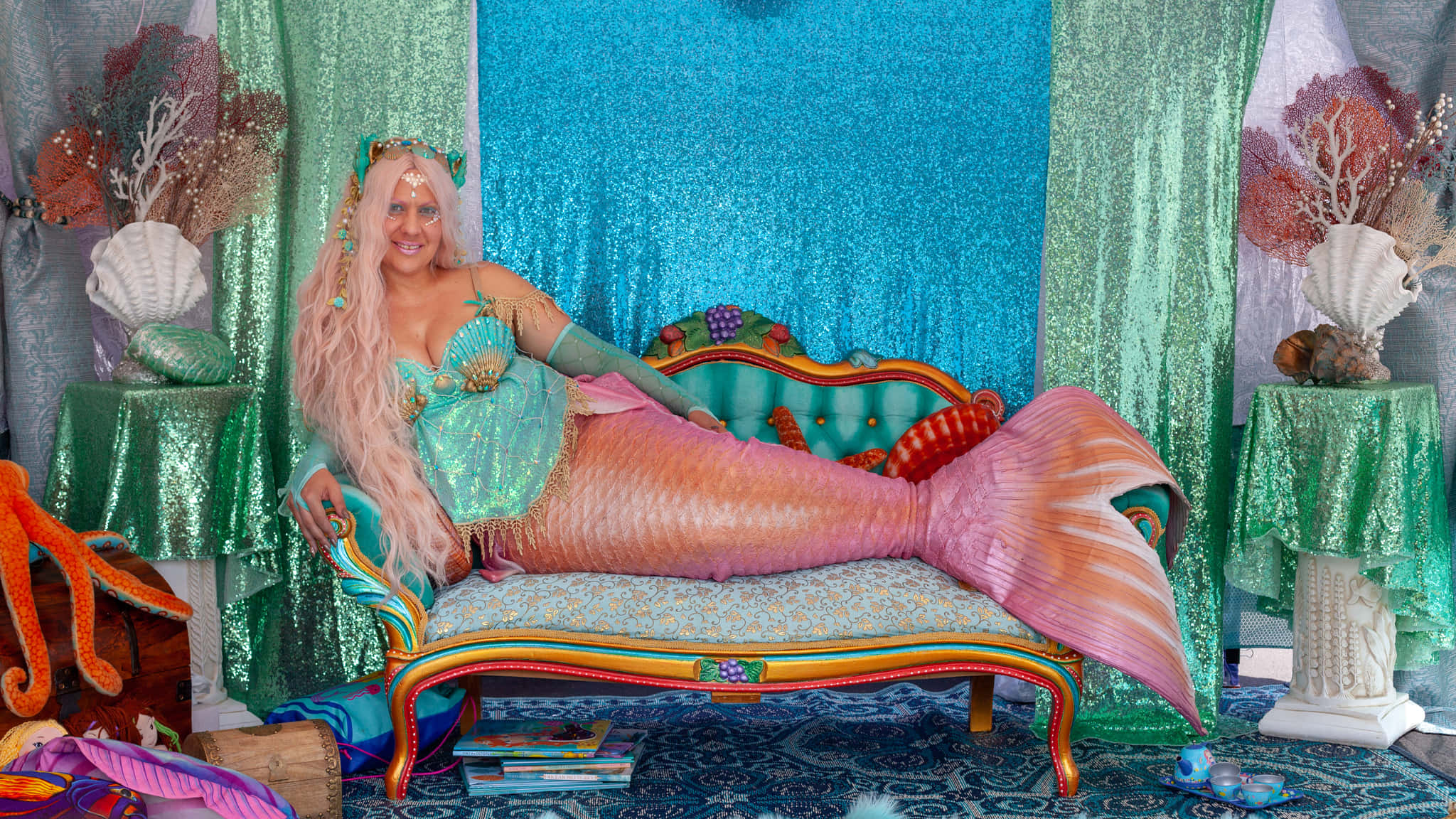 Real Life Mermaid 桌布 壁紙 — 一位微笑的美人魚，長長的粉紅色頭髮，躺在一張色彩繽紛、裝飾華麗的沙發上，背景是閃閃發光的藍綠色。
