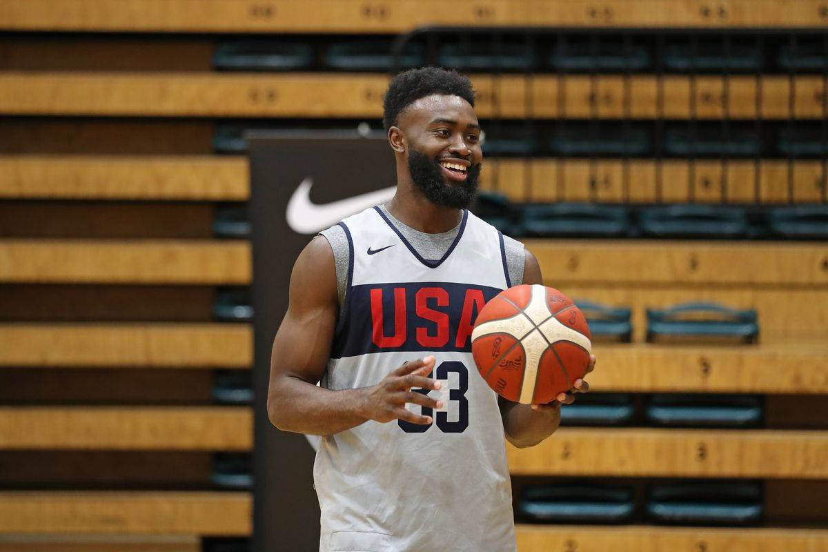 Jaylen Brown 桌布 壁紙 — 一位微笑的美國籃球運動員身穿白色球衣，在設有木製看台的體育館中手持一個紅白相間的球。