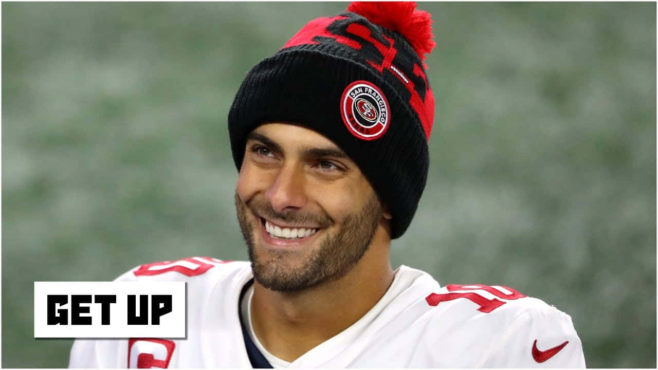 Jimmy Garoppolo 桌布 壁紙 — 一位微笑的美式足球員，戴著舊金山49人隊的毛帽和球衣，帶著燦爛的笑容望向鏡頭外。