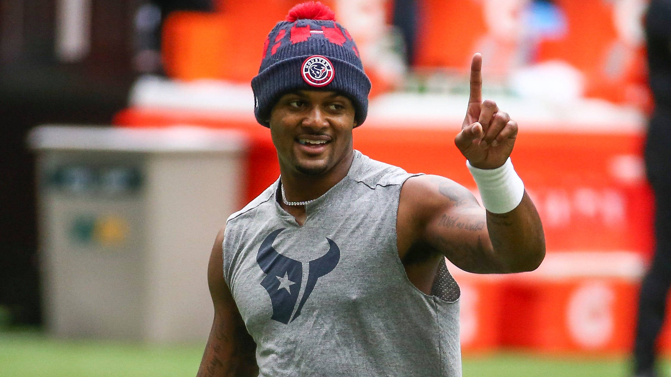 Deshaun Watson 桌布 壁紙 — 一位微笑的美式足球員戴著休士頓德州人隊的毛帽和無袖球衣，在訓練場上指向天空。