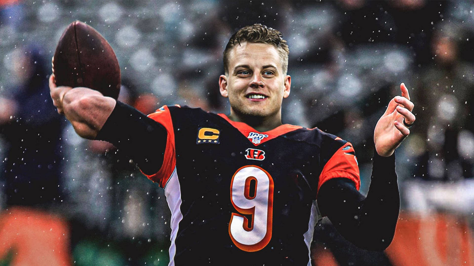 Joe Burrow 桌布 壁紙 — 一位微笑的美式足球四分衛身穿黑色和橙色9號球衣，在雨中拿著球並向上指。