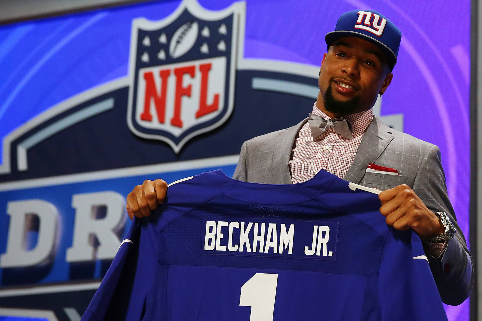 Odell 桌布 壁紙 — 一位微笑的美式足球運動員身穿西裝，在NFL選秀活動中舉起他的藍色紐約巨人隊球衣，上面印有「BECKHAM JR.」和1號。