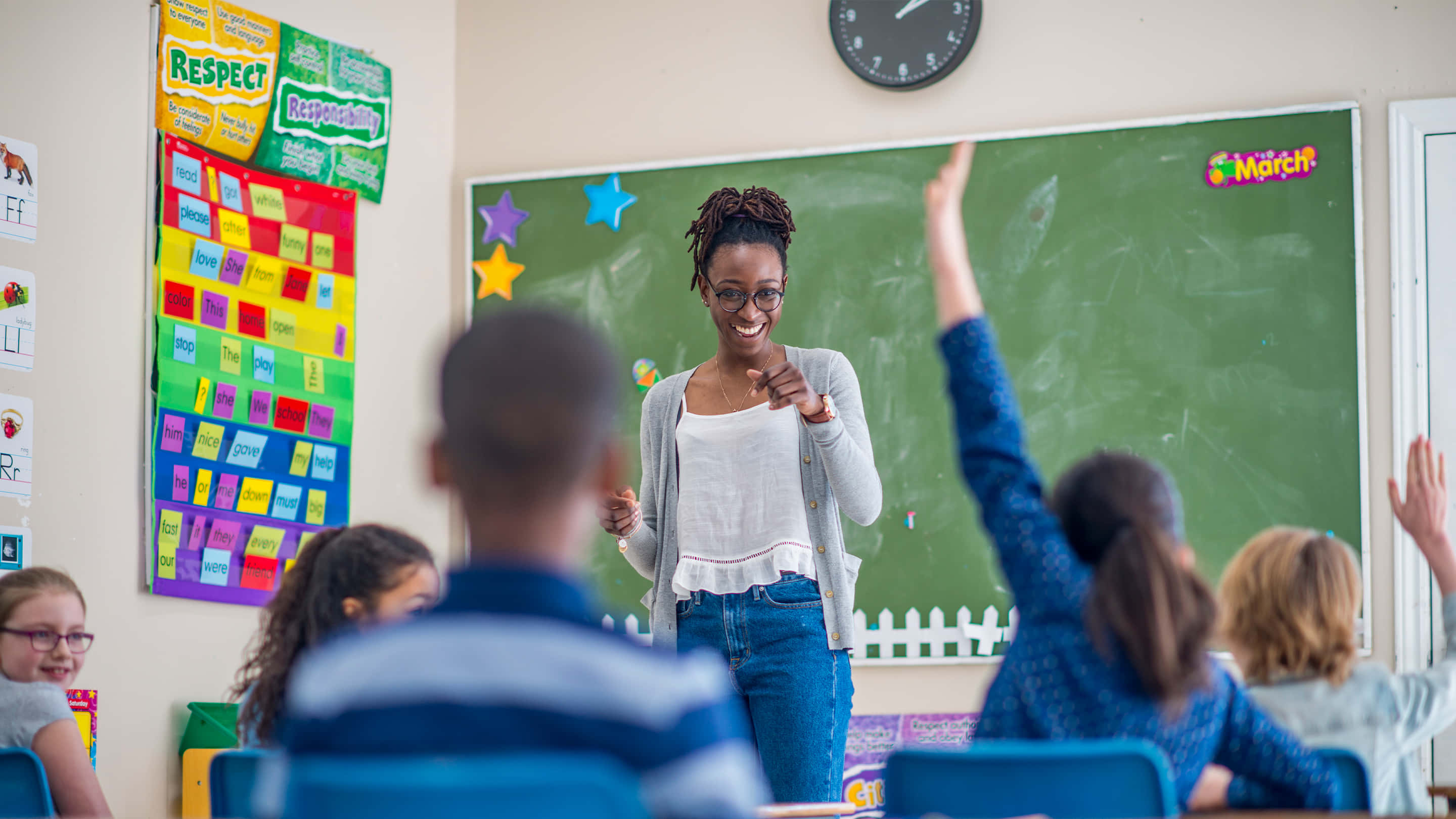 Classroom 桌布 壁紙 — 一位微笑的老師在充滿活力的教室中與學生互動，孩子們在課堂上熱切地舉手。