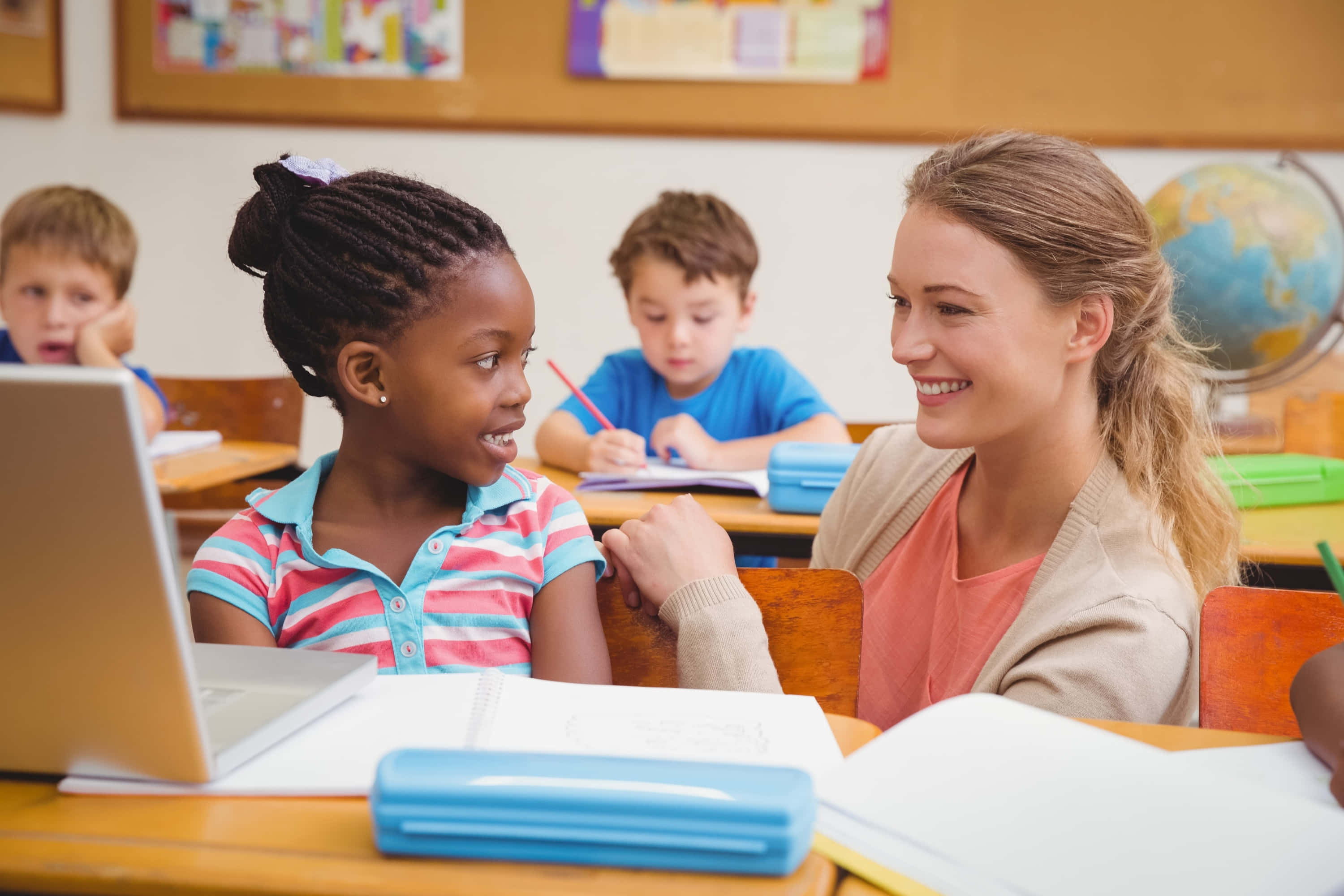 Learning 桌布 壁紙 — 一位微笑的老師在教室中與一位小女孩互動，其他學生在書桌上學習，背景有一個地球儀。