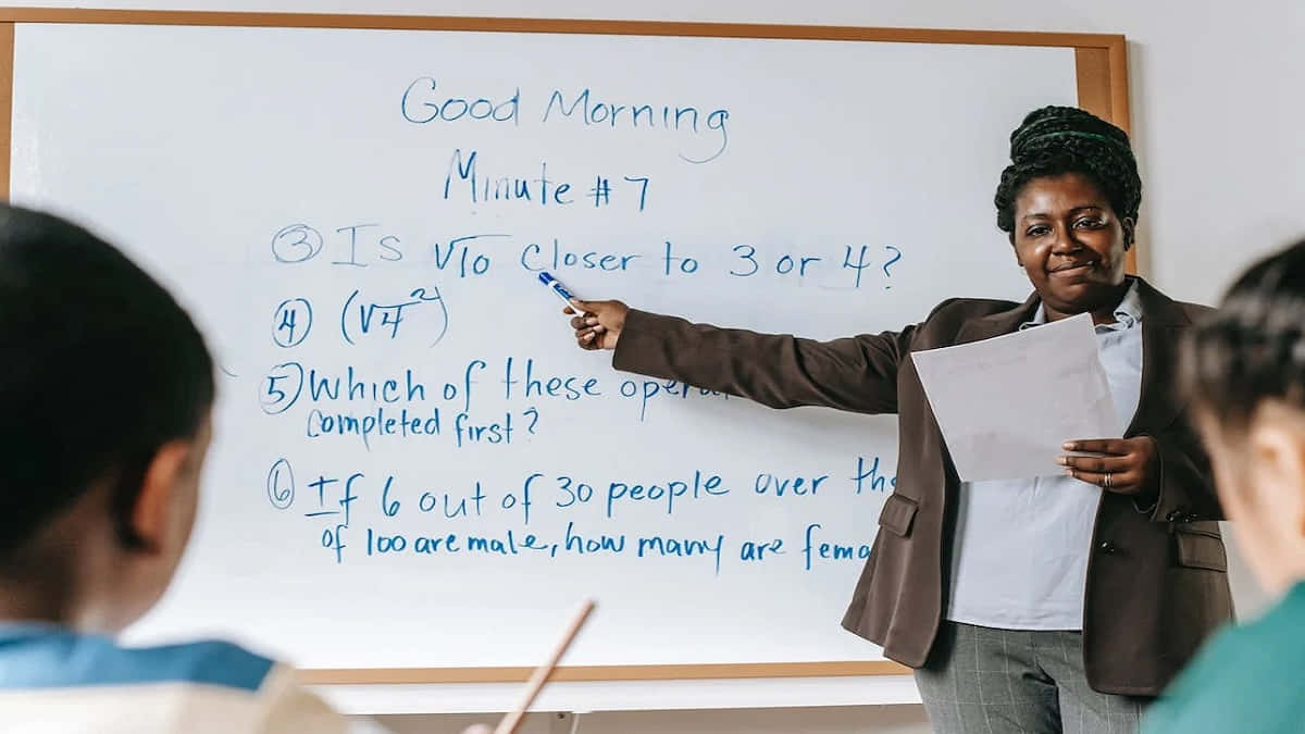 Learning 桌布 壁紙 — 一位微笑的老師指著白板上的數學問題，學生們在教室裡面對著她。
