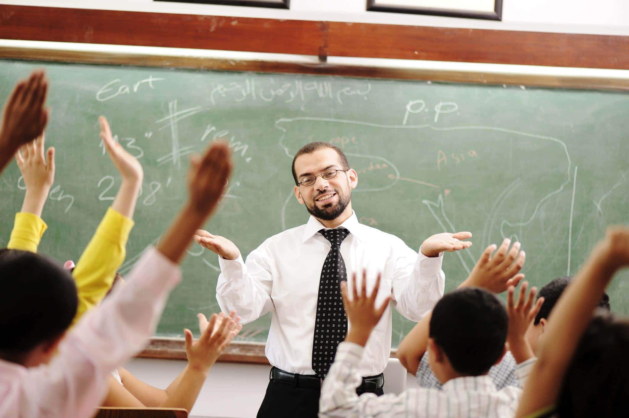 teachers 桌布 壁紙 — 一位微笑的老師熱情地做手勢，學生們在一個充滿活力的教室裡熱情舉手，身後有一塊黑板地圖。