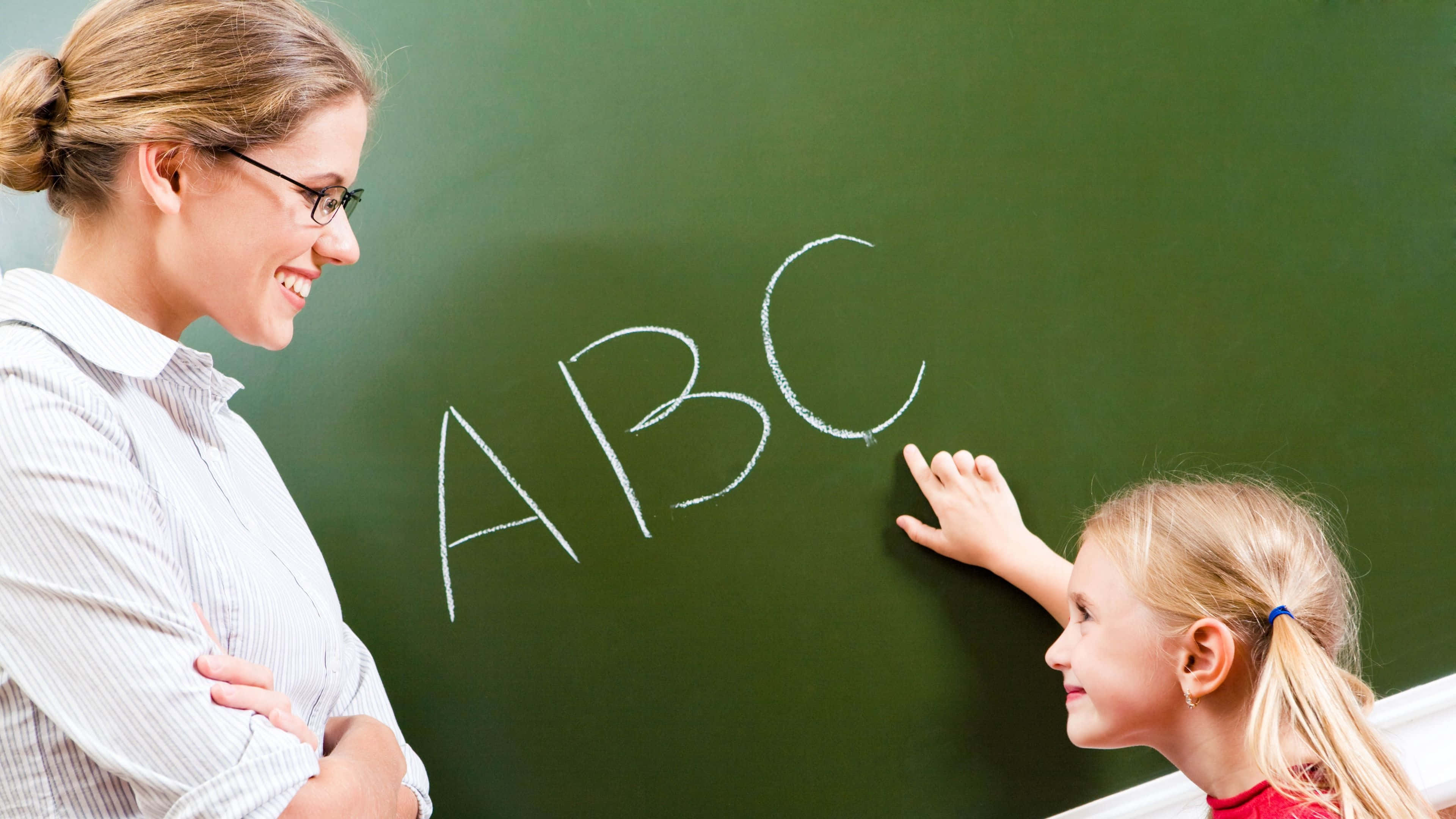 teachers 桌布 壁紙 — 一位微笑的老師看著一個小女孩指著綠色黑板上的粉筆字母ABC，在明亮的教室裡。