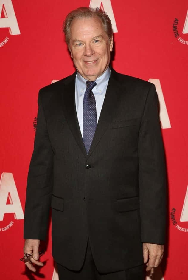 Michael Mckean 桌布 壁紙 — 一位微笑的老年男士，身穿黑色西裝和圖案領帶，自信地站在帶有白色標誌的鮮紅色背景前。