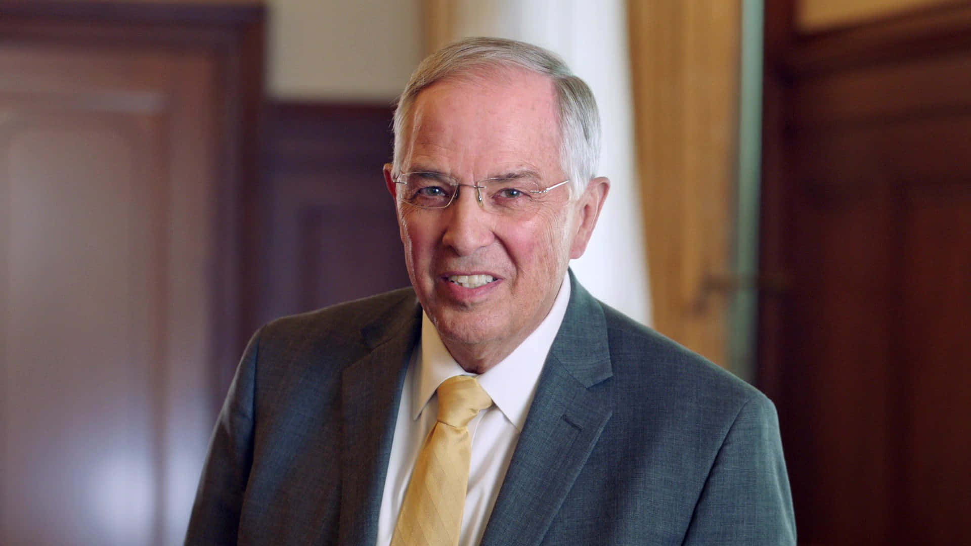 Mormon 桌布 壁紙 — 一位微笑的老年男士身穿灰色西裝和黃色領帶，站在一個有木質鑲板和柔和燈光的房間裡。