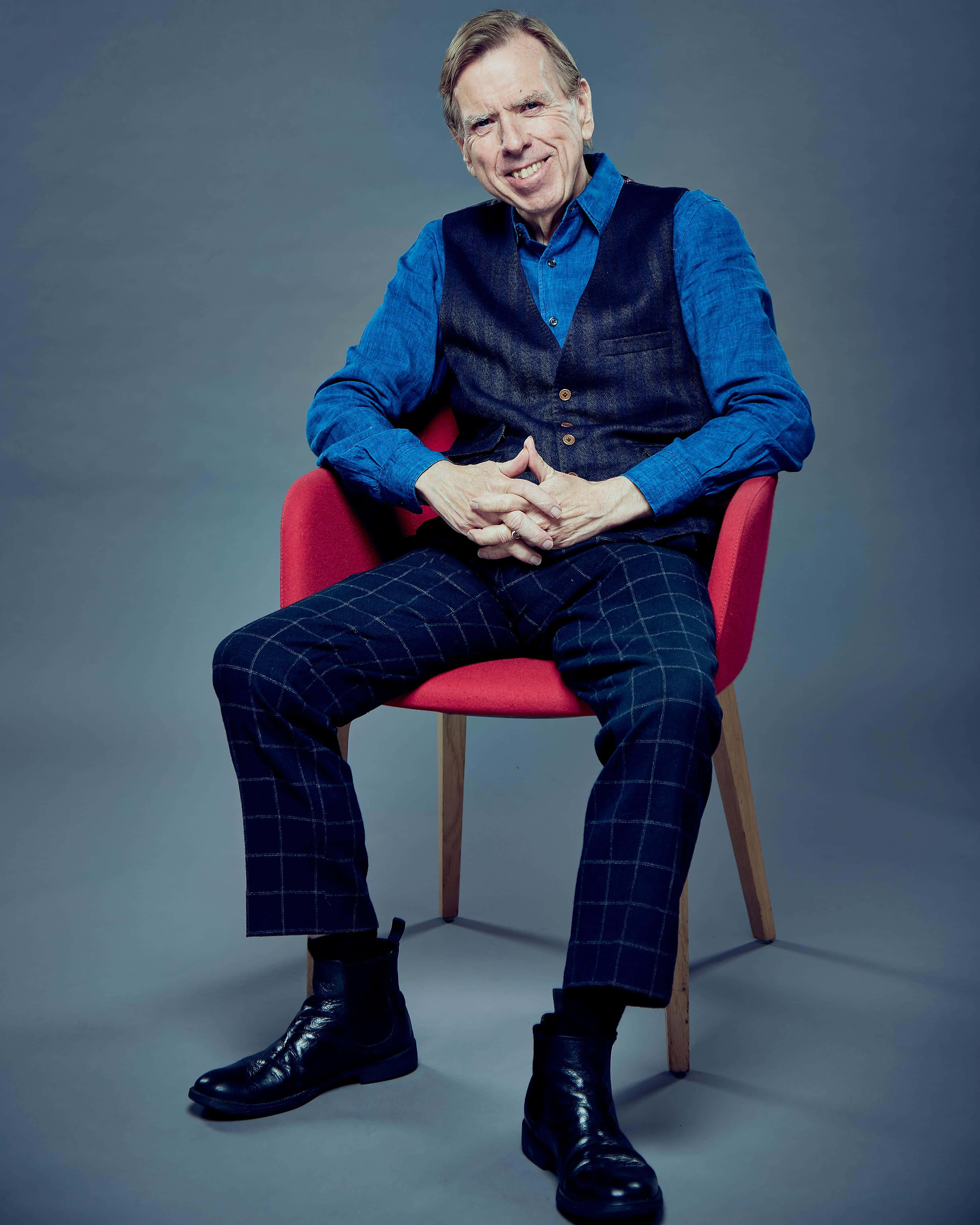 Timothy Spall 桌布 壁紙 — 一位微笑的老年男子，身穿藍色襯衫和格子褲，自信地坐在一張鮮紅色的椅子上，背景是柔和的灰色攝影棚。