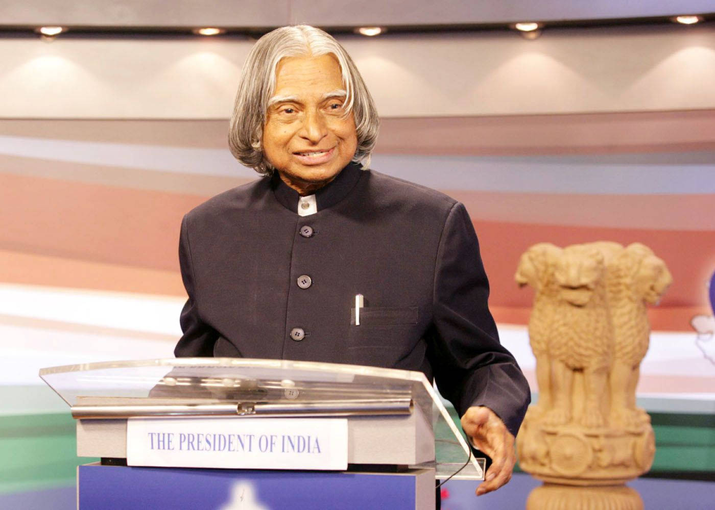 smiling 桌布 壁紙 — 一位微笑的老年男子身穿深色庫爾塔，站在標有「THE PRESIDENT OF INDIA」的講台後面，旁邊有一個石製的阿育王獅子柱頭。