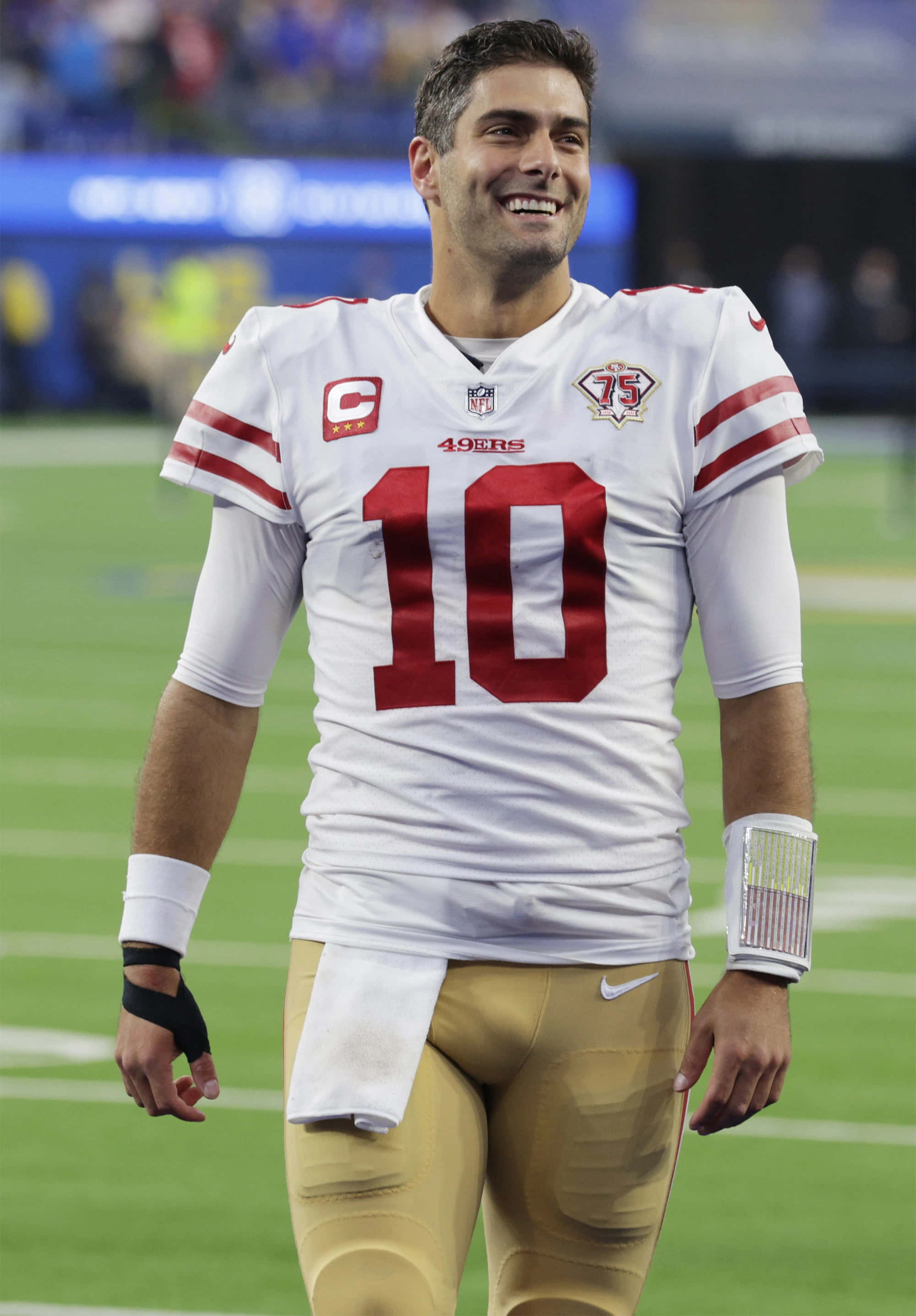 Jimmy Garoppolo 桌布 壁紙 — 一位微笑的舊金山49人四分衛，身穿白色和紅色10號球衣，在綠色足球場上行走。