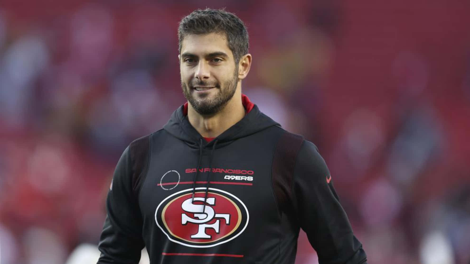 Jimmy Garoppolo 桌布 壁紙 — 一位微笑的舊金山49人球員穿著黑色球隊連帽衫，自信地站在模糊的球場上。