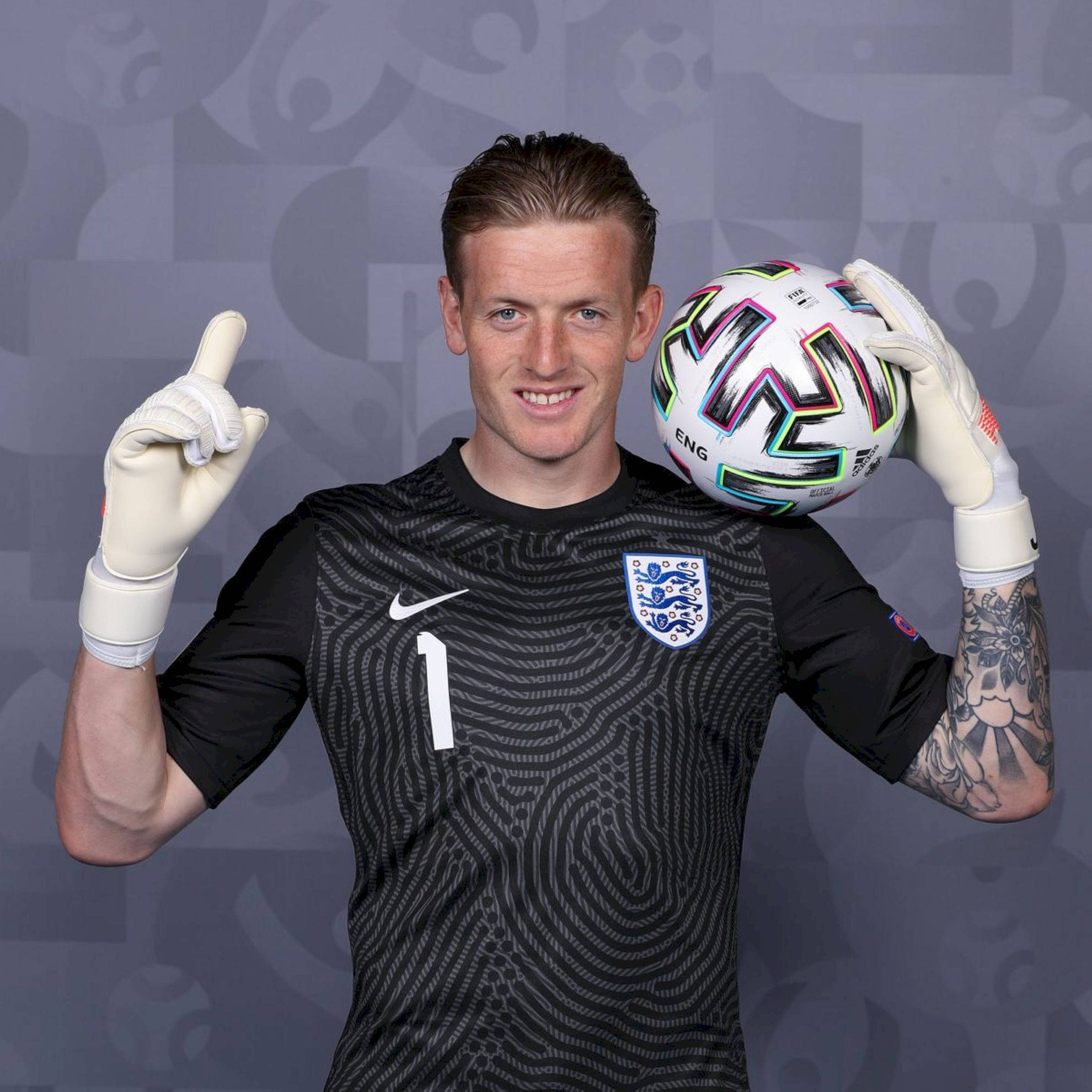 Football Players 桌布 壁紙 — 一位微笑的英格蘭守門員身穿黑色球衣，戴著手套的手拿著一個色彩繽紛的足球，同時向上指著。