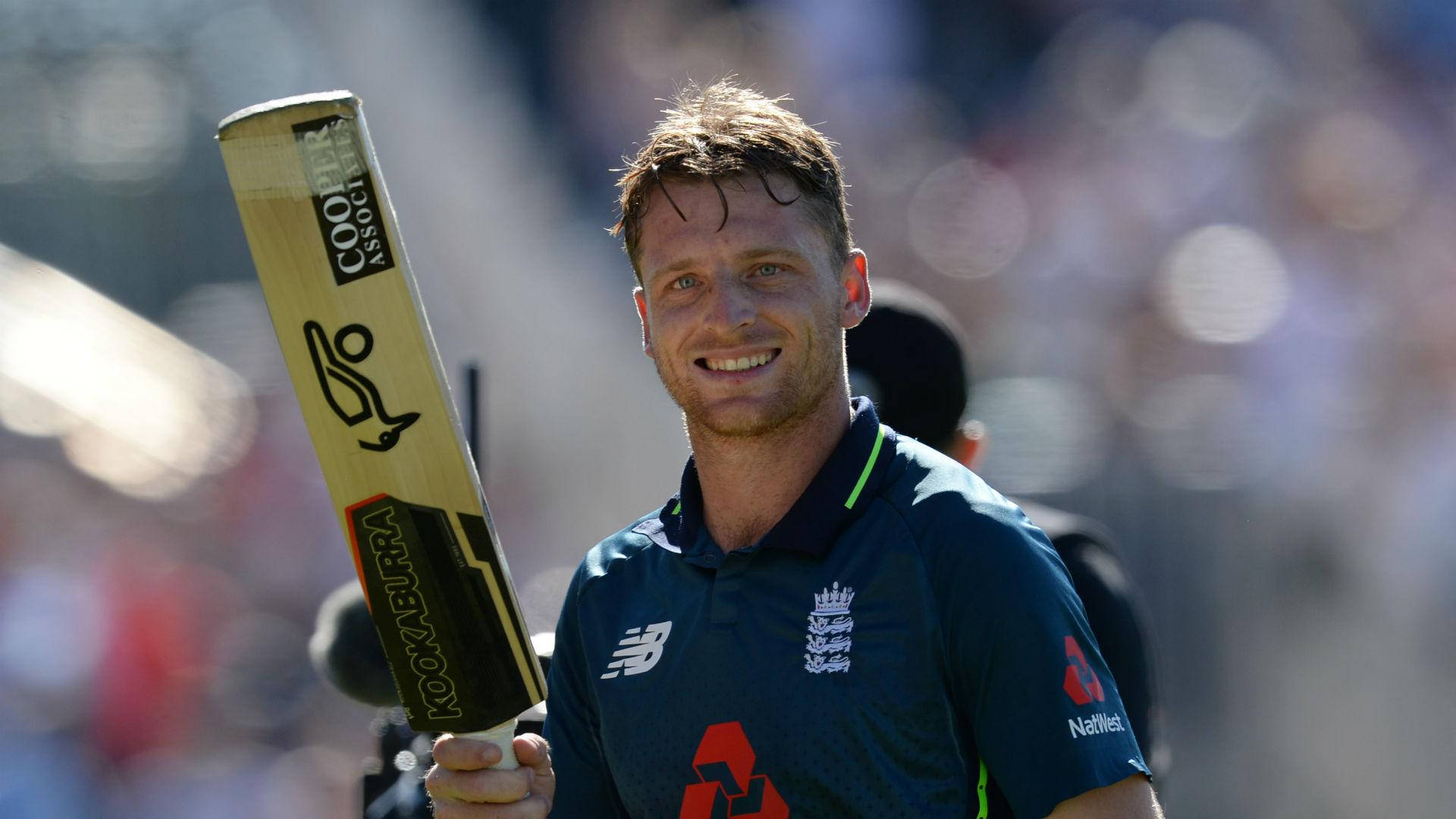 jos buttler 桌布 壁紙 — 一位微笑的英格蘭板球運動員，在明亮的陽光球場背景下，自豪地舉起一支 Kookaburra 球棒。
