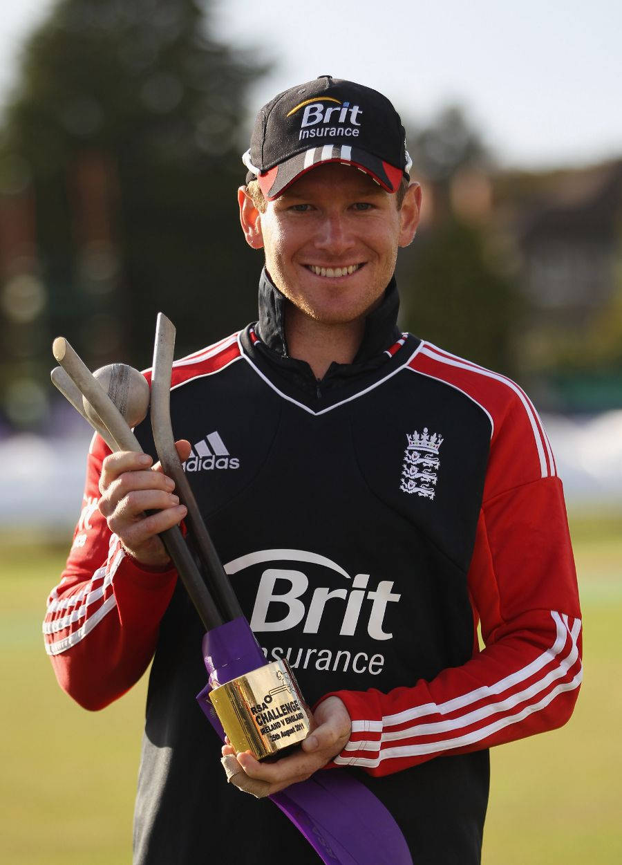 eoin morgan 桌布 壁紙 — 一位微笑的英格蘭板球運動員在明亮的日光下，於草地上手持金色獎盃和木製球棒。