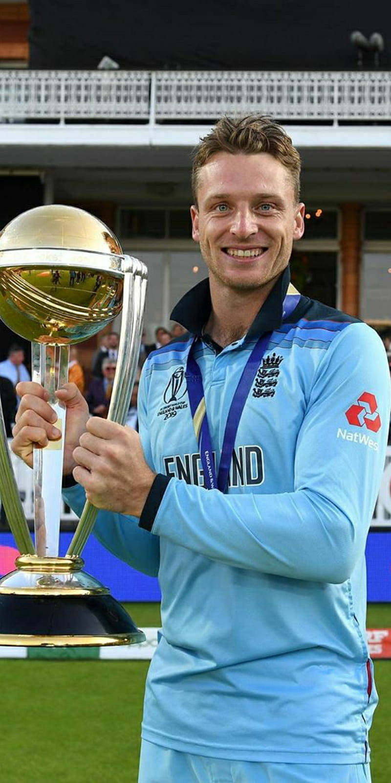 jos buttler 桌布 壁紙 — 一位微笑的英格蘭板球運動員身穿淺藍色制服，在綠色場地上自豪地高舉一個大型金色獎盃。