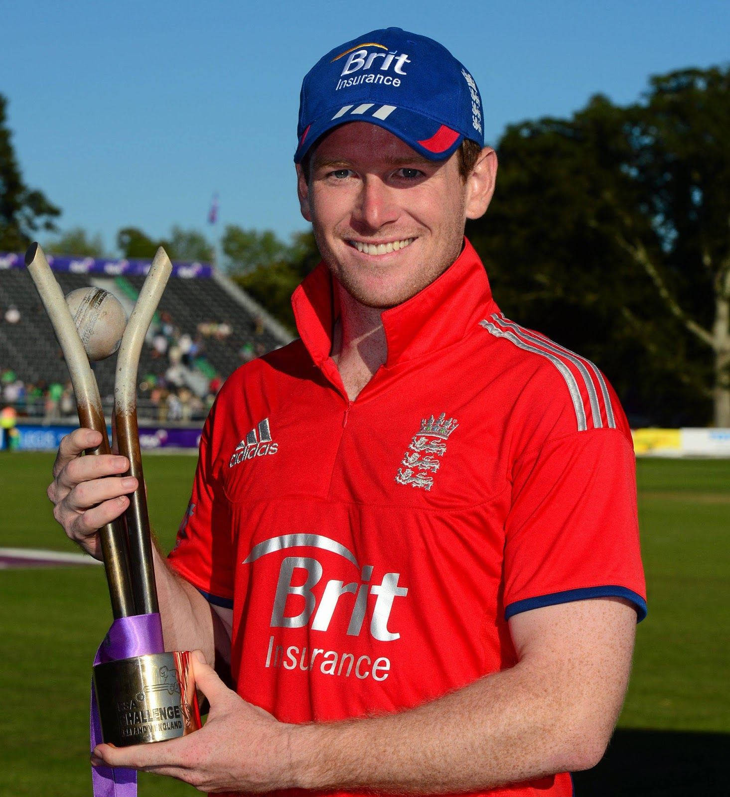 eoin morgan 桌布 壁紙 — 一位微笑的英格蘭板球運動員身穿紅色球衣，在陽光明媚的球場背景下，高舉頂部有板球的獎盃。