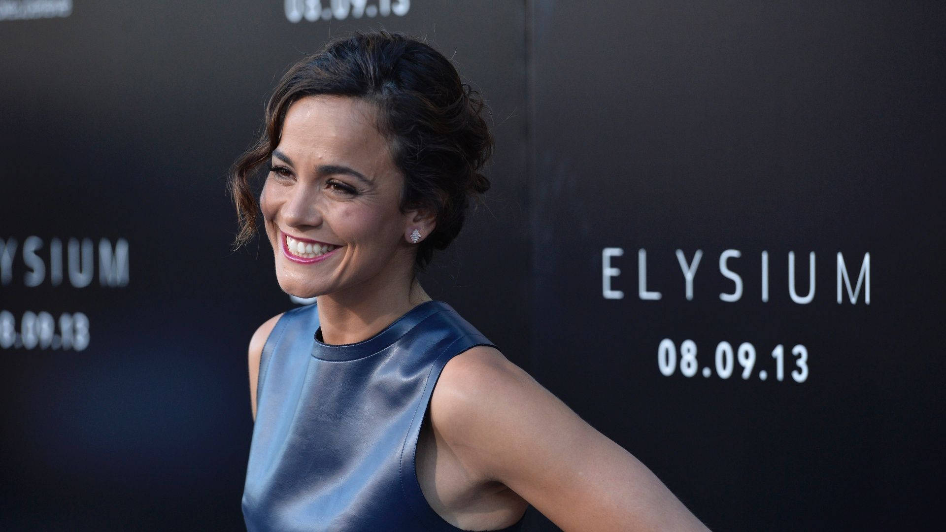 Celebrity 桌布 壁紙 — 一位微笑的藍裙女士在電影首映會上擺姿勢，身後有「Elysium」和日期文字。