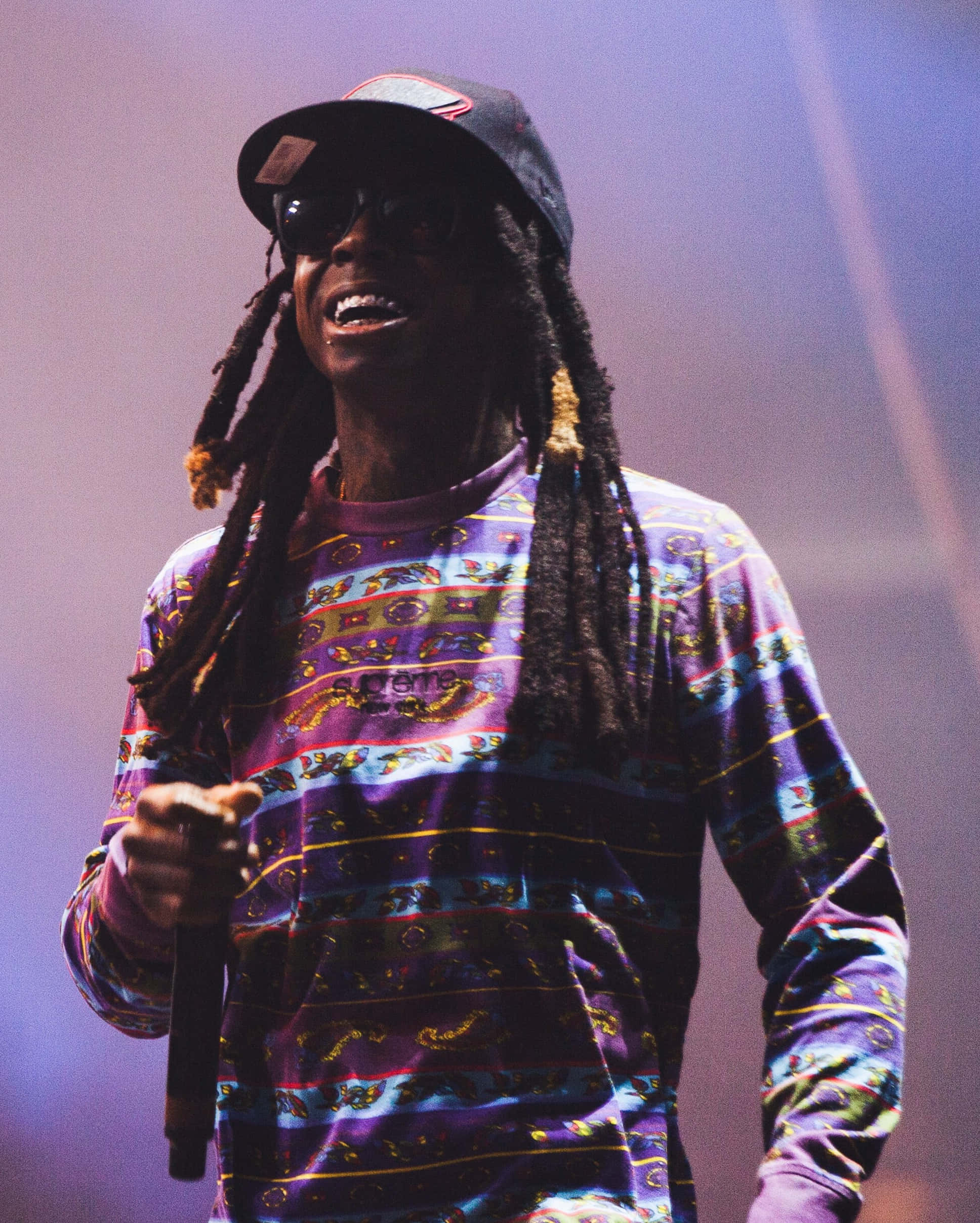 2 Chainz 桌布 壁紙 — 一位微笑的表演者，留著辮子頭、戴著太陽眼鏡，在紫色舞台燈光下對著麥克風唱歌，穿著色彩繽紛的圖案襯衫。