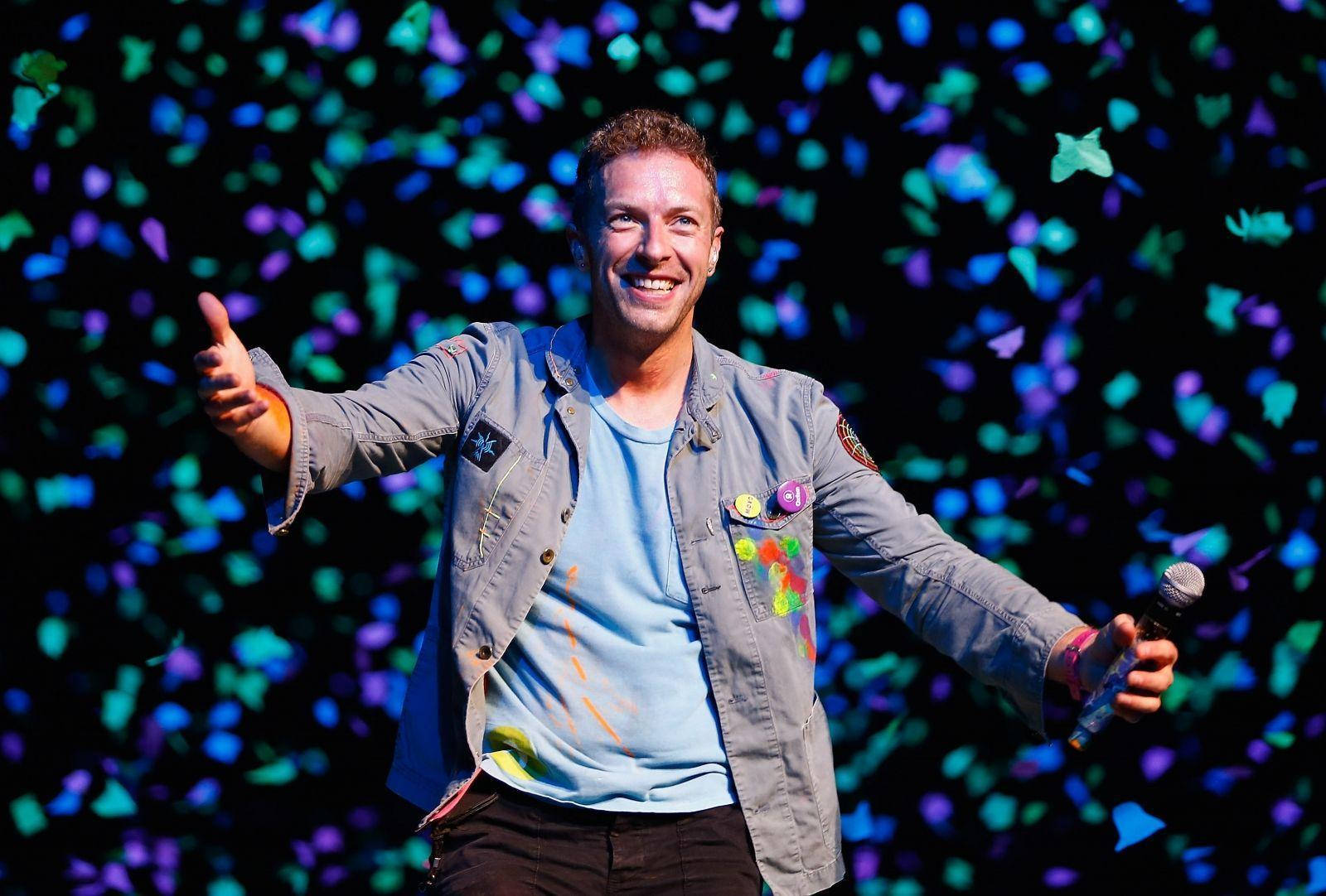 Chris Martin 桌布 壁紙 — 一位微笑的表演者在舞台上張開雙臂，置身於飄落的彩色紙屑中，背景昏暗。