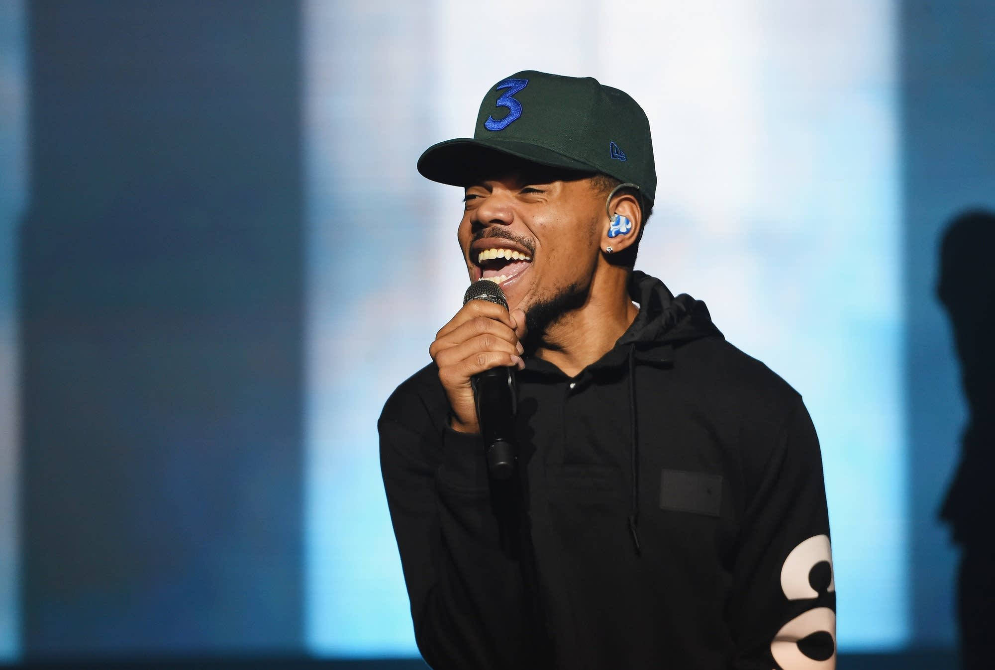 chance 桌布 壁紙 — 一位微笑的表演者戴著綠色帽子和黑色連帽衫，在明亮的舞台燈光下對著麥克風唱歌。