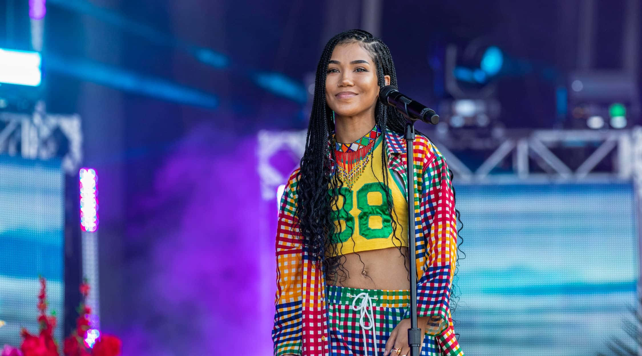 jhene aiko 桌布 壁紙 — 一位微笑的表演者穿著色彩繽紛的格子服裝，在閃爍燈光下自信地站在充滿活力的舞台上。