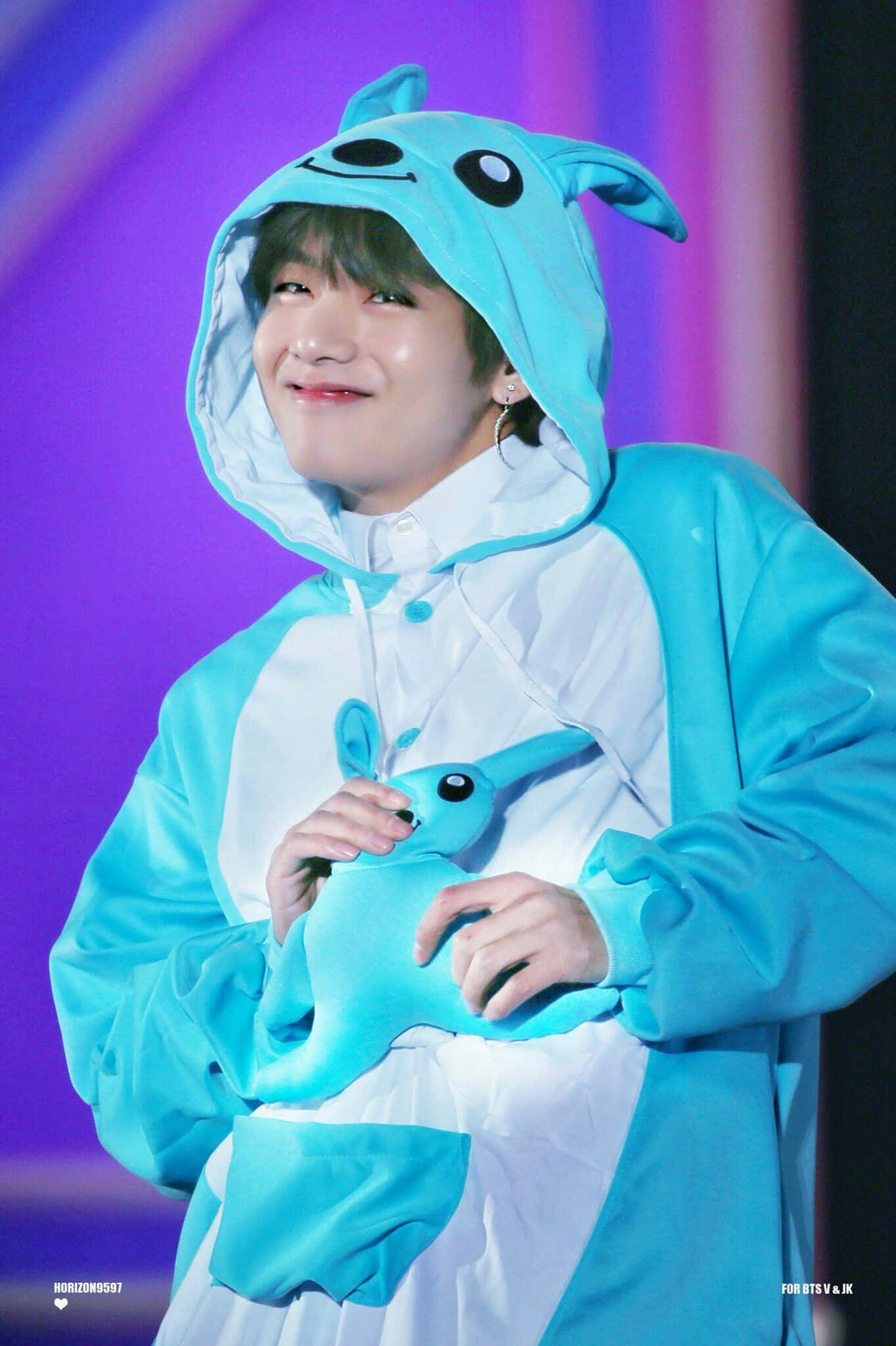 Taehyung Cute 桌布 壁紙 — 一位微笑的表演者穿著藍白兔子連身衣，在充滿活力的紫色舞台背景下抱著一個相配的毛絨玩具。