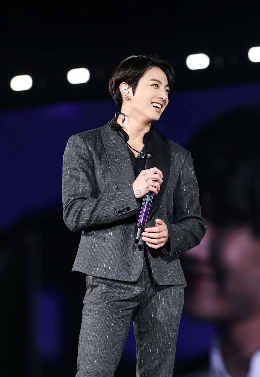 Jungkook 桌布 壁紙 — 一位微笑的表演者穿著閃亮的灰色西裝，在黑暗背景下明亮的舞台燈光下握著麥克風。