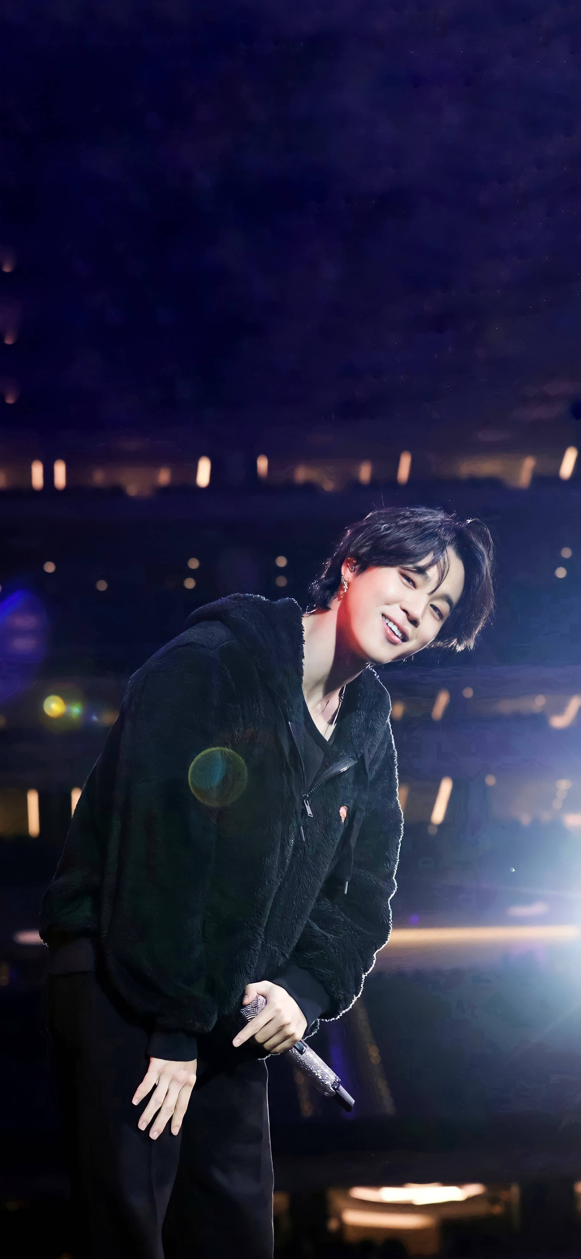 Bts Jimin 桌布 壁紙 — 一位微笑的表演者穿著黑色連帽衫，在舞台燈光下拿著麥克風，背景是模糊的觀眾。