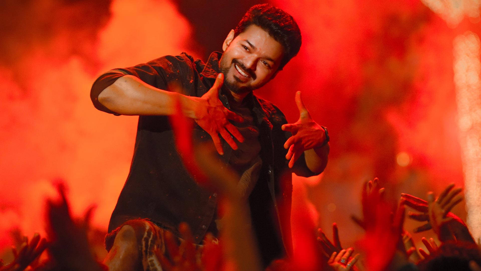 Bollywood HD 桌布 壁紙 — 一位微笑的表演者身穿黑色襯衫，在火紅舞台燈光下，於高舉雙手的群眾面前充滿活力地跳舞。