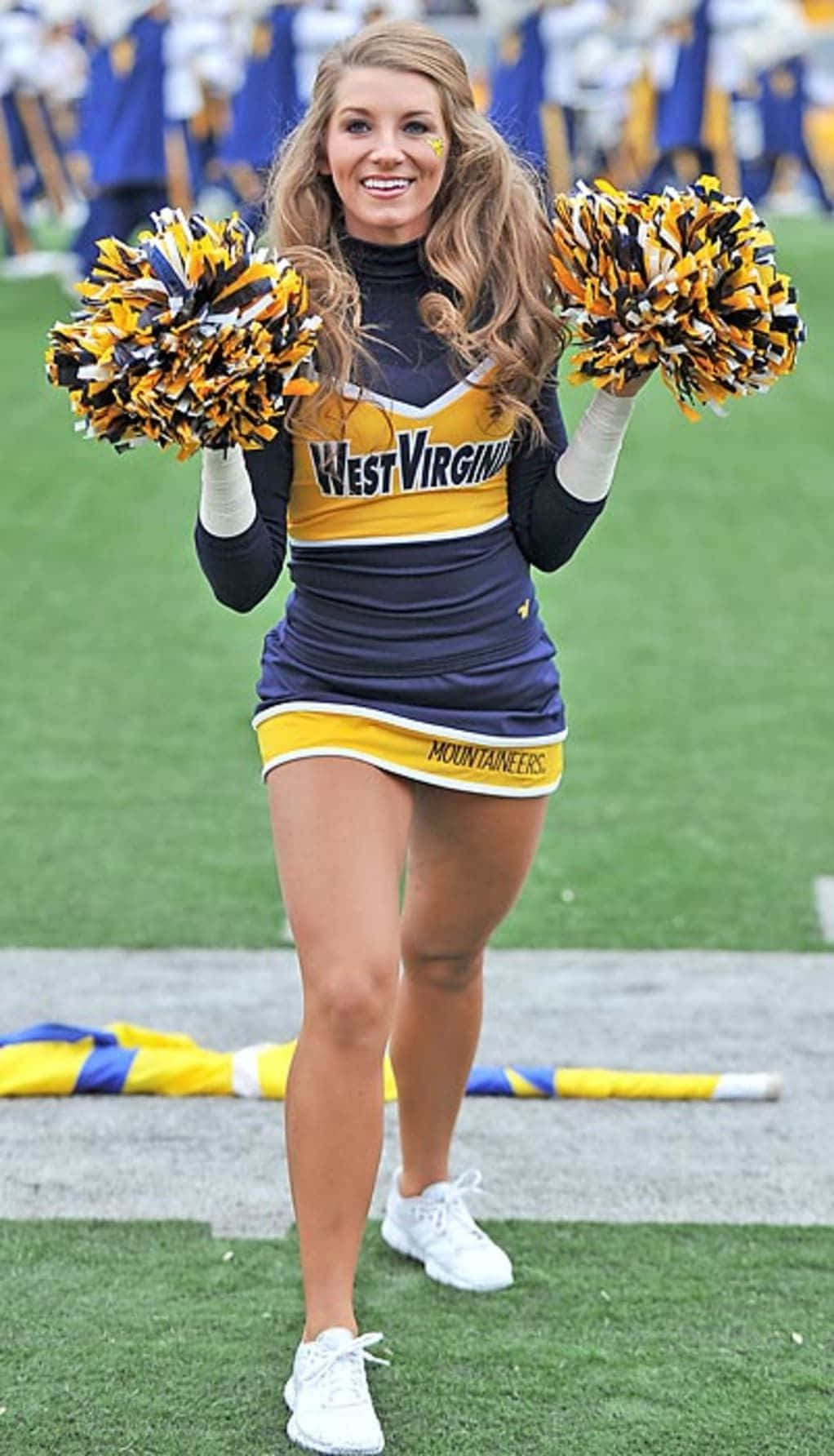 Cheering 桌布 壁紙 — 一位微笑的西維吉尼亞登山者啦啦隊員身穿海軍藍和金色制服，在足球場上手持彩球。