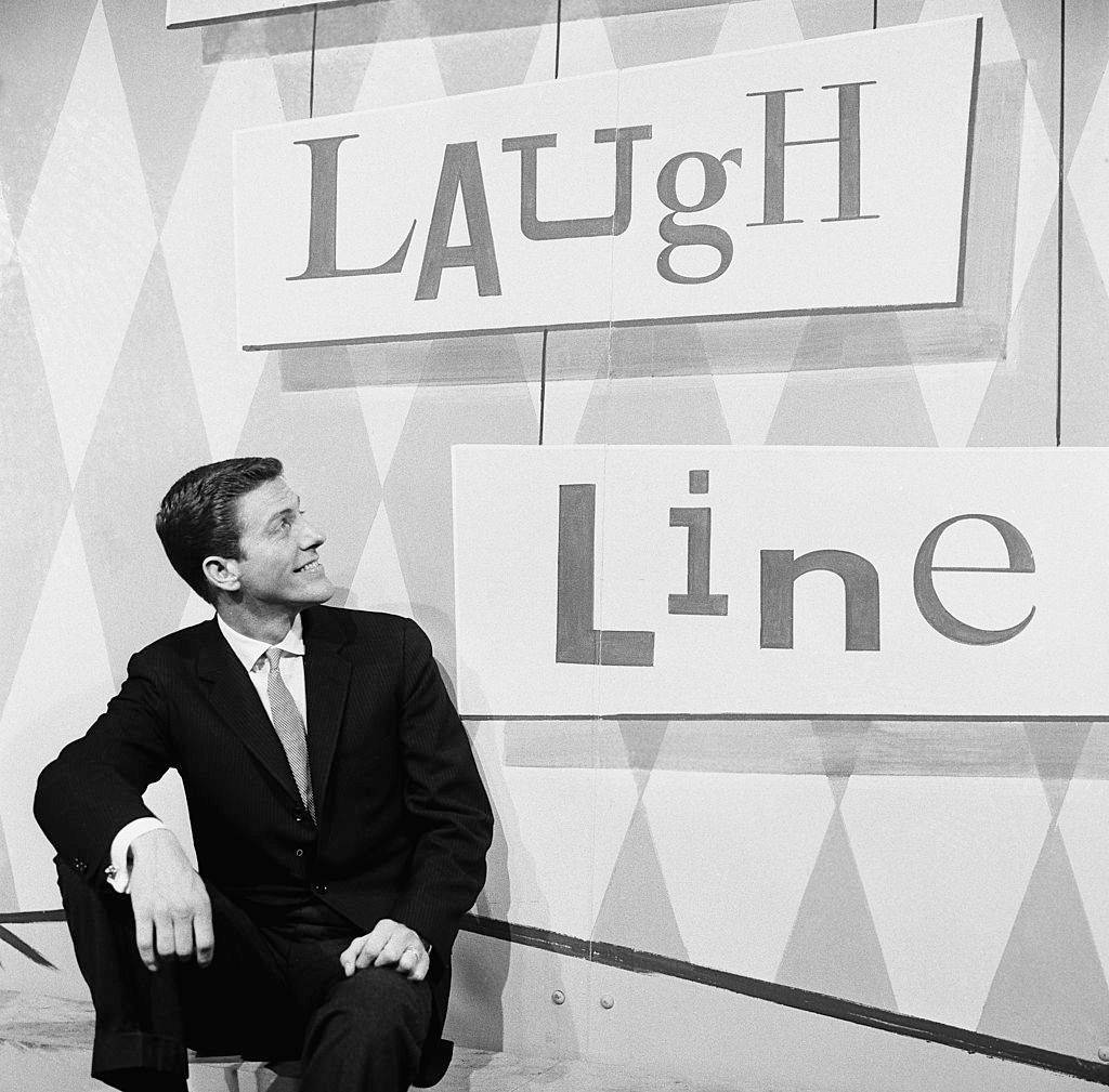 美國 桌布 壁紙 — 一位微笑的西裝男士坐在幾何牆前，牆上有 'LAUGH Line' 標誌，呈現復古黑白風格。
