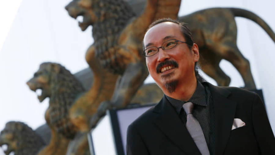 satoshi kon 桌布 壁紙 — 一位微笑的西裝男子在明亮的日光下站在一排青銅獅子雕像前。