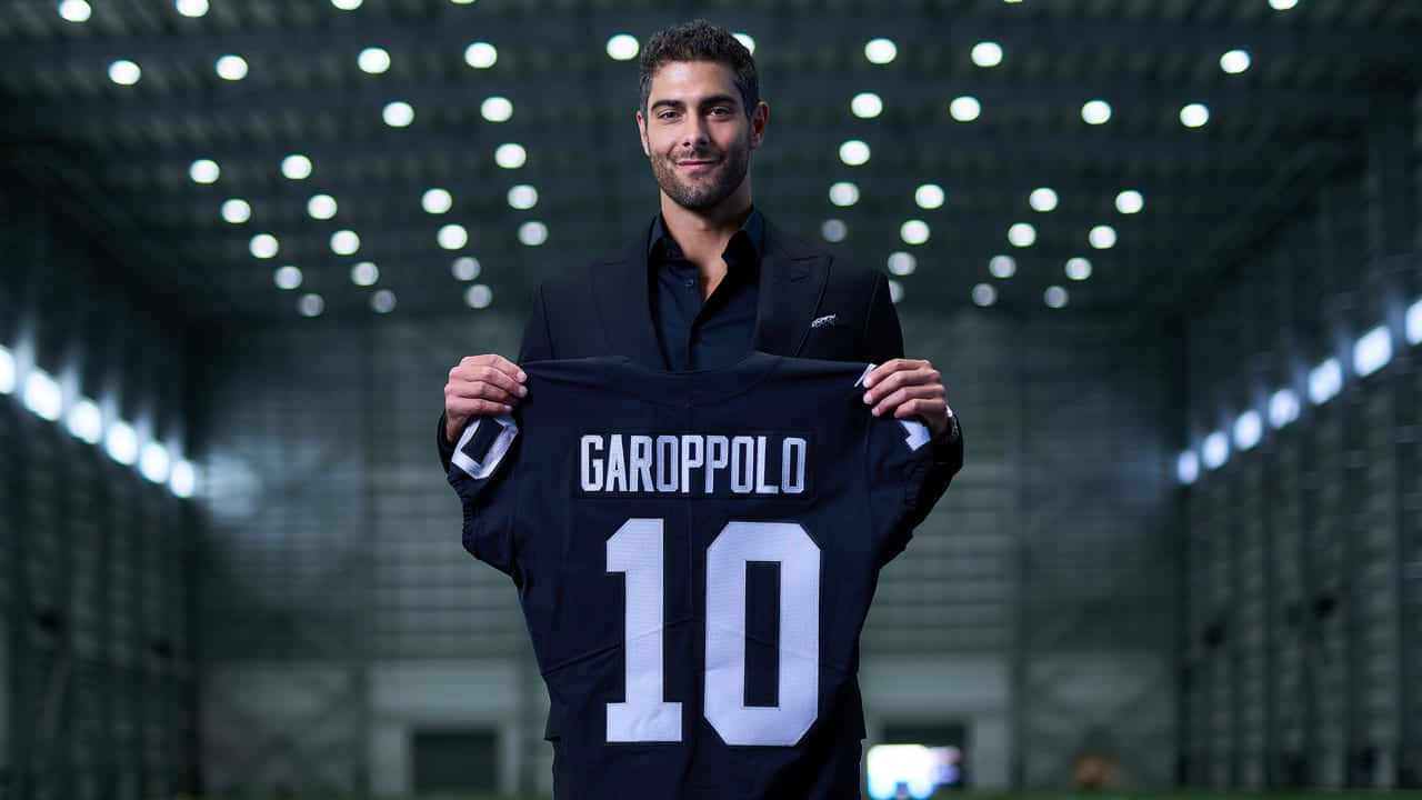 Jimmy Garoppolo 桌布 壁紙 — 一位微笑的西裝男子在明亮的體育場燈光下，舉起一件印有白色 'GAROPPOLO 10' 字樣的黑色足球球衣。