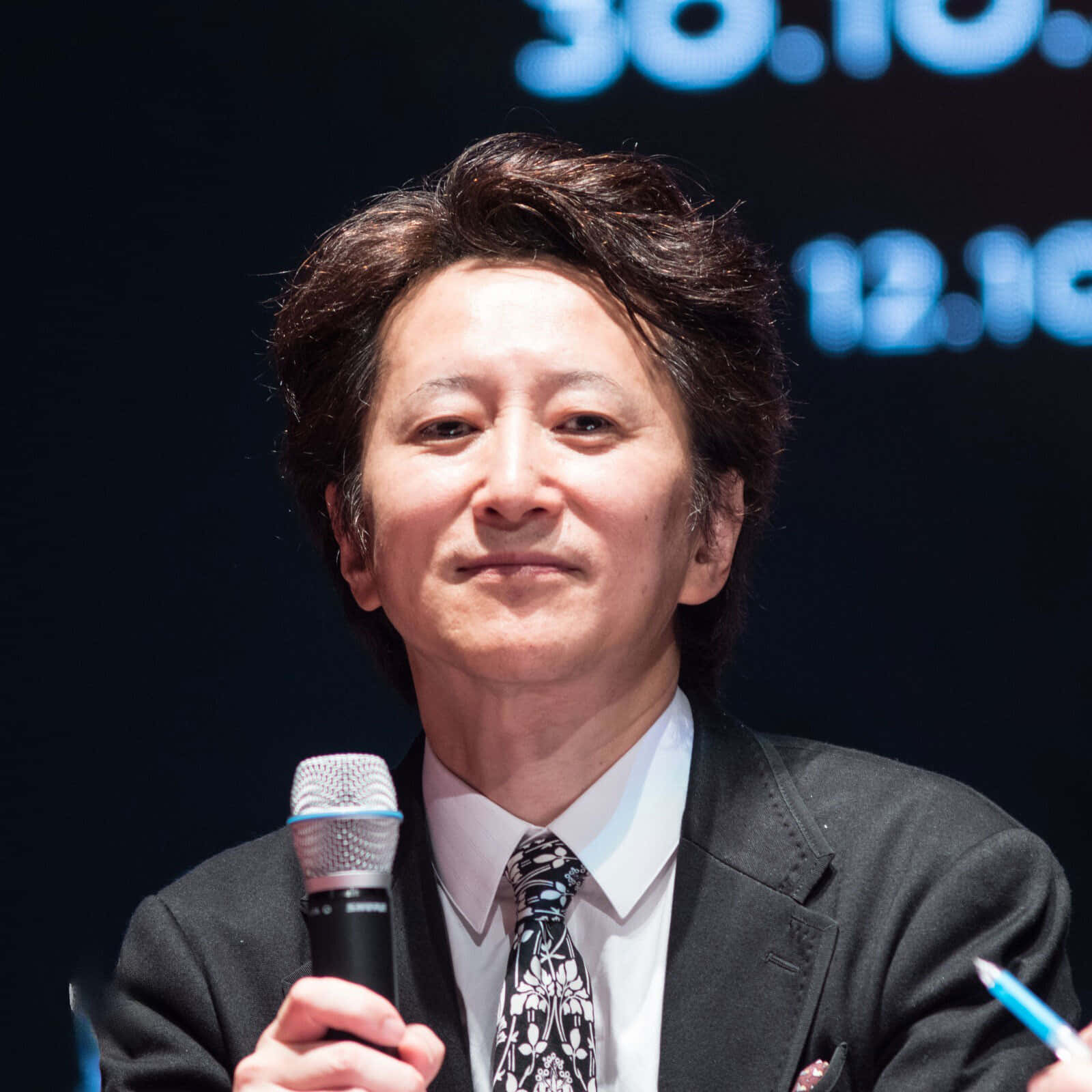 Hirohiko Araki 桌布 壁紙 — 一位微笑的西裝男子在黑暗背景下手持麥克風，背景有發光的藍色數字。