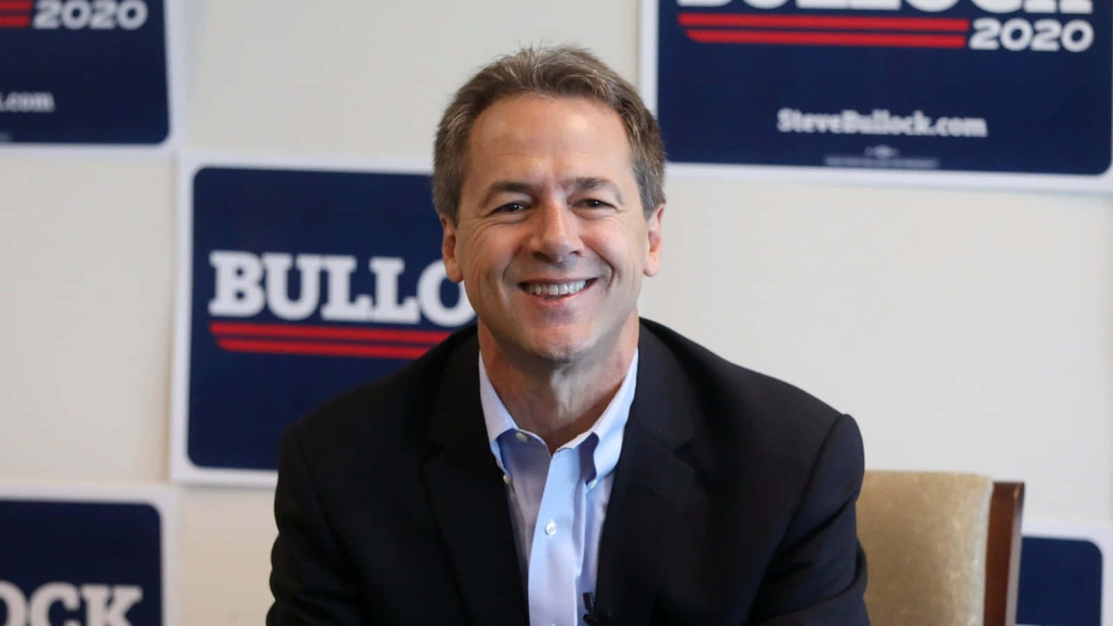 政治 桌布 壁紙 — 一位微笑的西裝男子坐在Steve Bullock 2020競選標誌前的新聞發布會場景。