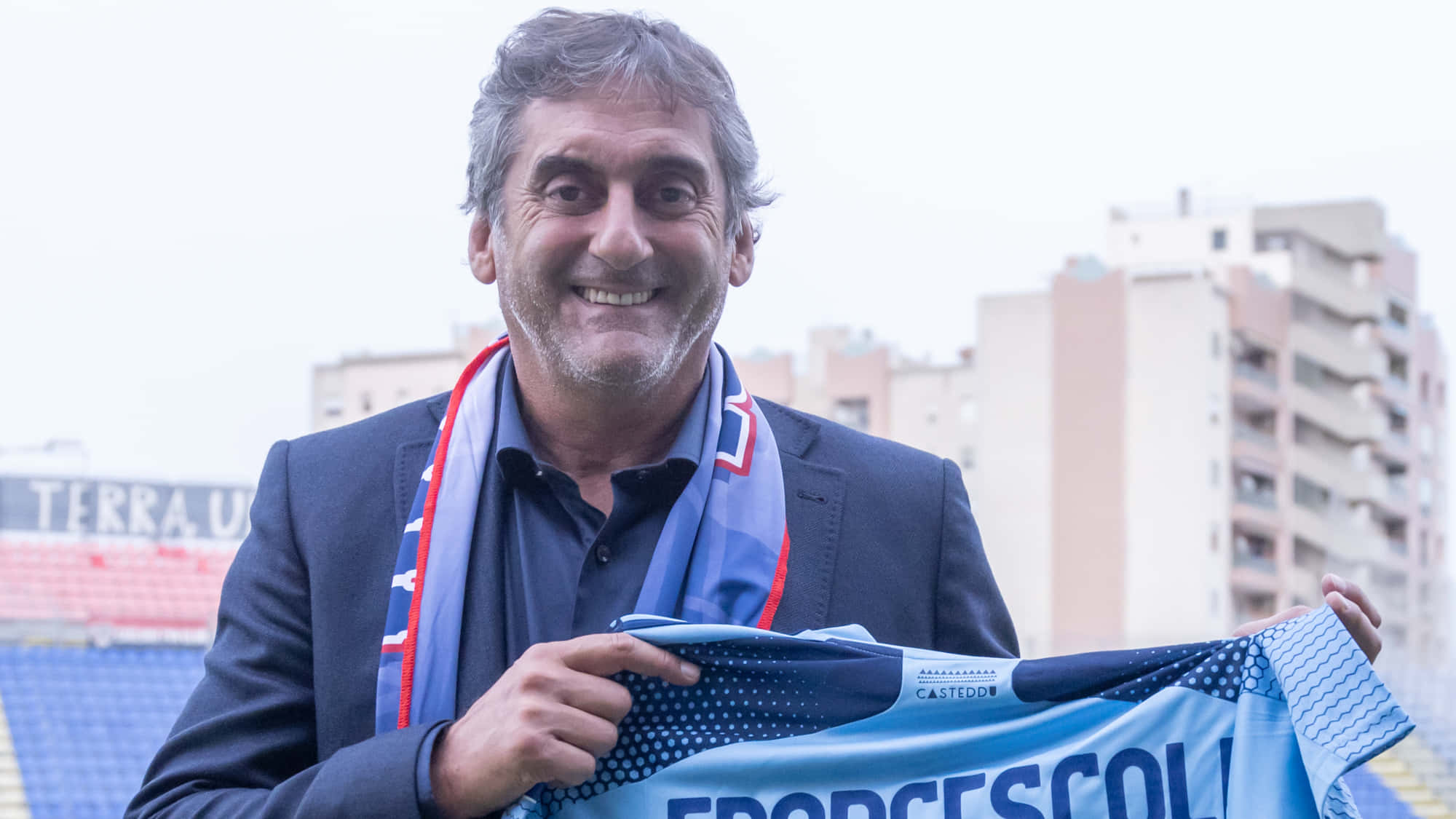 足球 桌布 壁紙 — 一位微笑的西裝男子手持印有 'FRANCESCOLI' 的淺藍色足球球衣，站在一個有紅色座位和城市建築背景的體育場內。