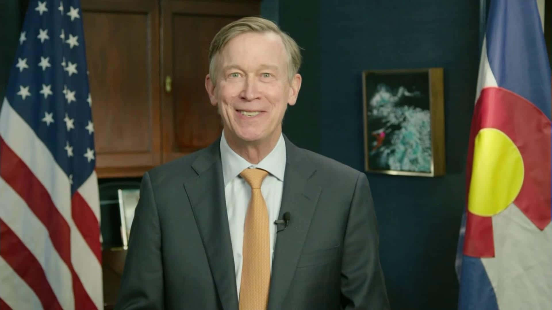 American 桌布 壁紙 — 一位微笑的西裝男子站在美國國旗和科羅拉多州旗之間，處於正式辦公室環境。