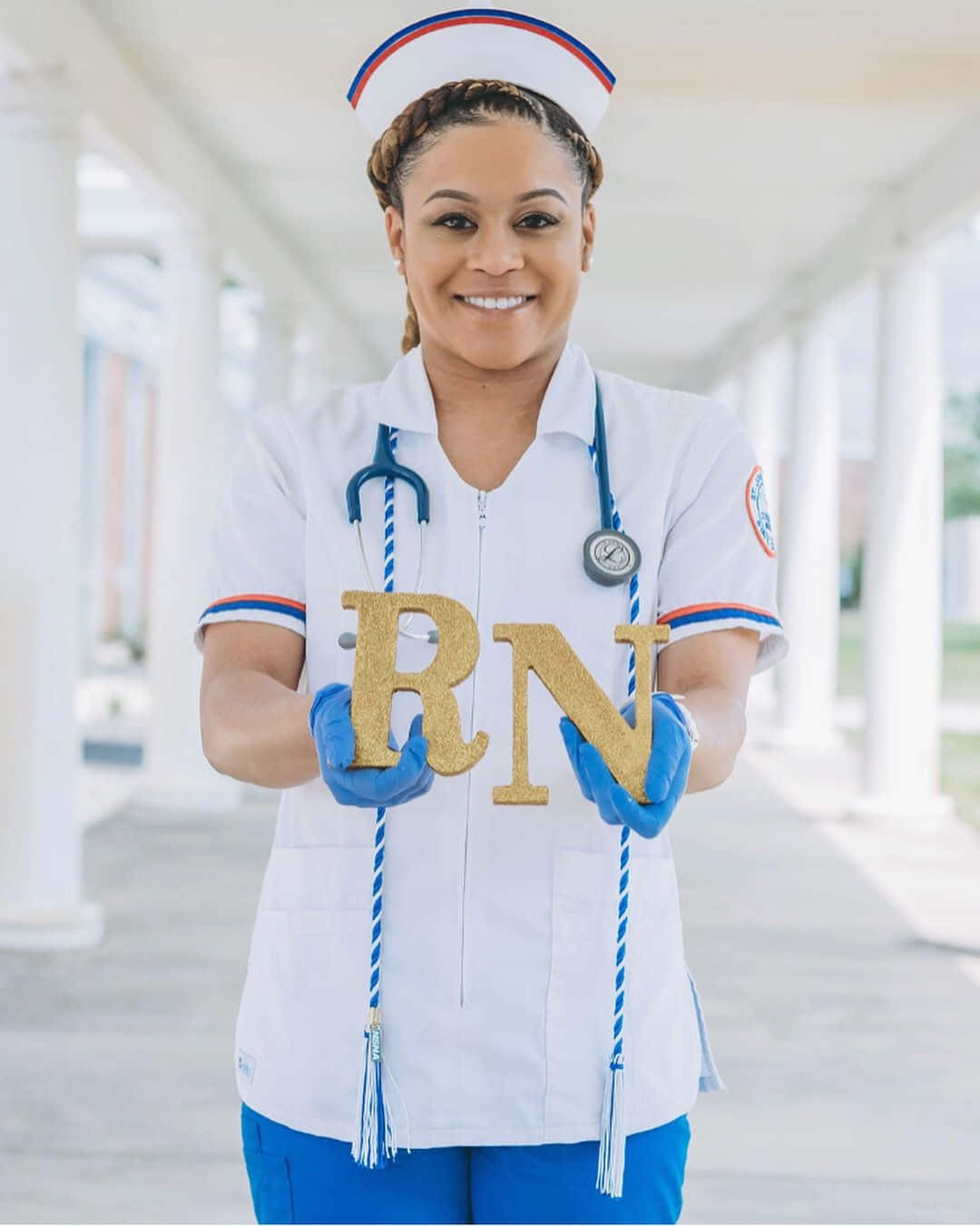 Nurse Graduation 桌布 壁紙 — 一位微笑的護士穿著白色制服和藍色手套，在明亮的日光下自豪地舉起閃閃發光的金色RN字母。