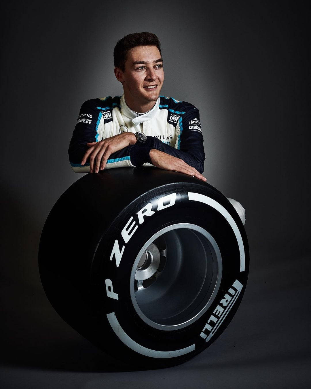 george russell 桌布 壁紙 — 一位微笑的賽車手穿著團隊制服，在黑暗的攝影棚背景下，隨意地靠著一個巨大的 Pirelli P Zero 賽車輪胎。