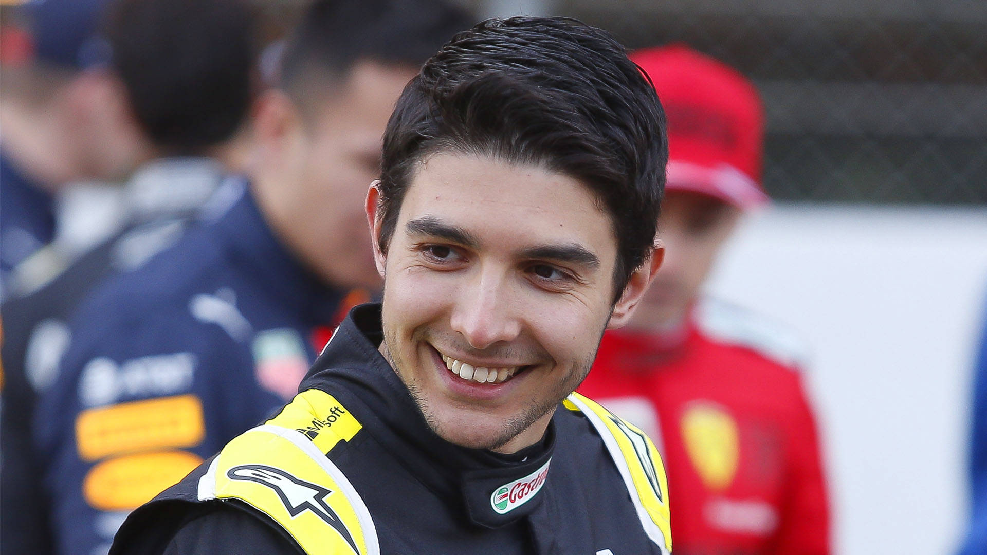 F1 桌布 壁紙 — 一位微笑的賽車手穿著帶有贊助商標誌的黑黃色車隊外套，在賽車活動中看向鏡頭。