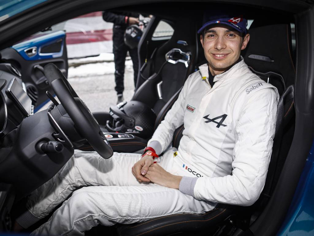 F1 桌布 壁紙 — 一位微笑的賽車手穿著白色賽車服，自信地坐在藍色賽車內，可見方向盤和頭盔。