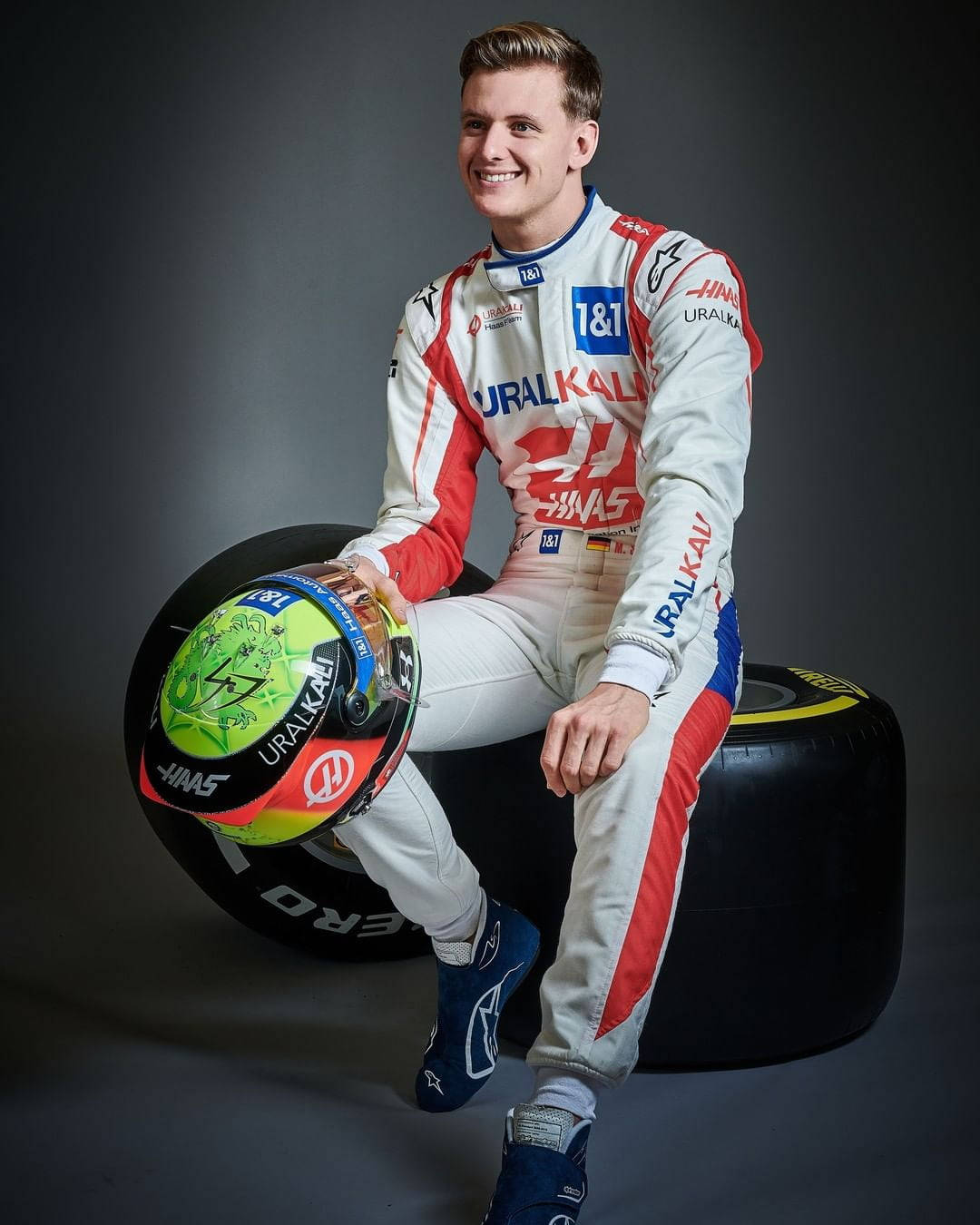 mick schumacher 桌布 壁紙 — 一位微笑的賽車手身穿白色、紅色和藍色的賽車服，隨意地坐在黑色輪胎上，手持色彩繽紛的頭盔。