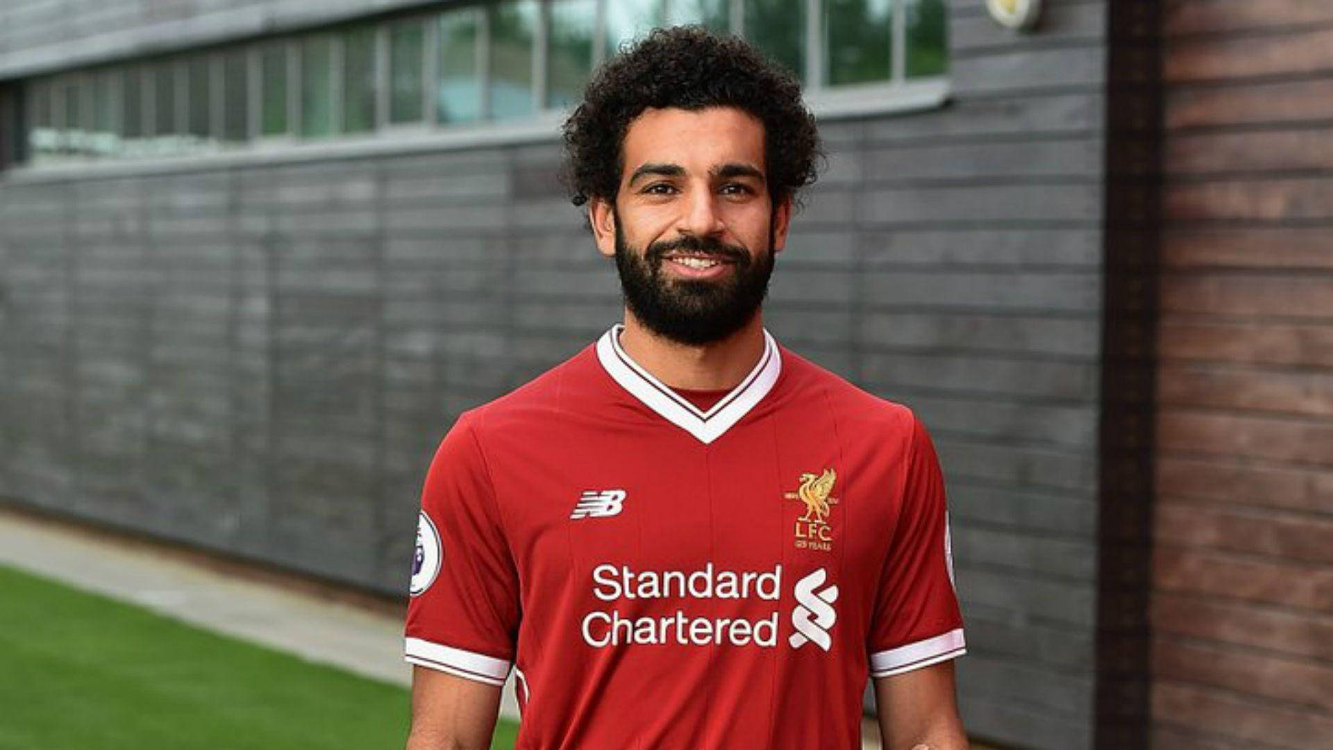 Salah 桌布 壁紙 — 一位微笑的足球員，捲髮和鬍鬚，身穿紅色利物浦FC球衣，背景是現代木質和灰色牆壁。