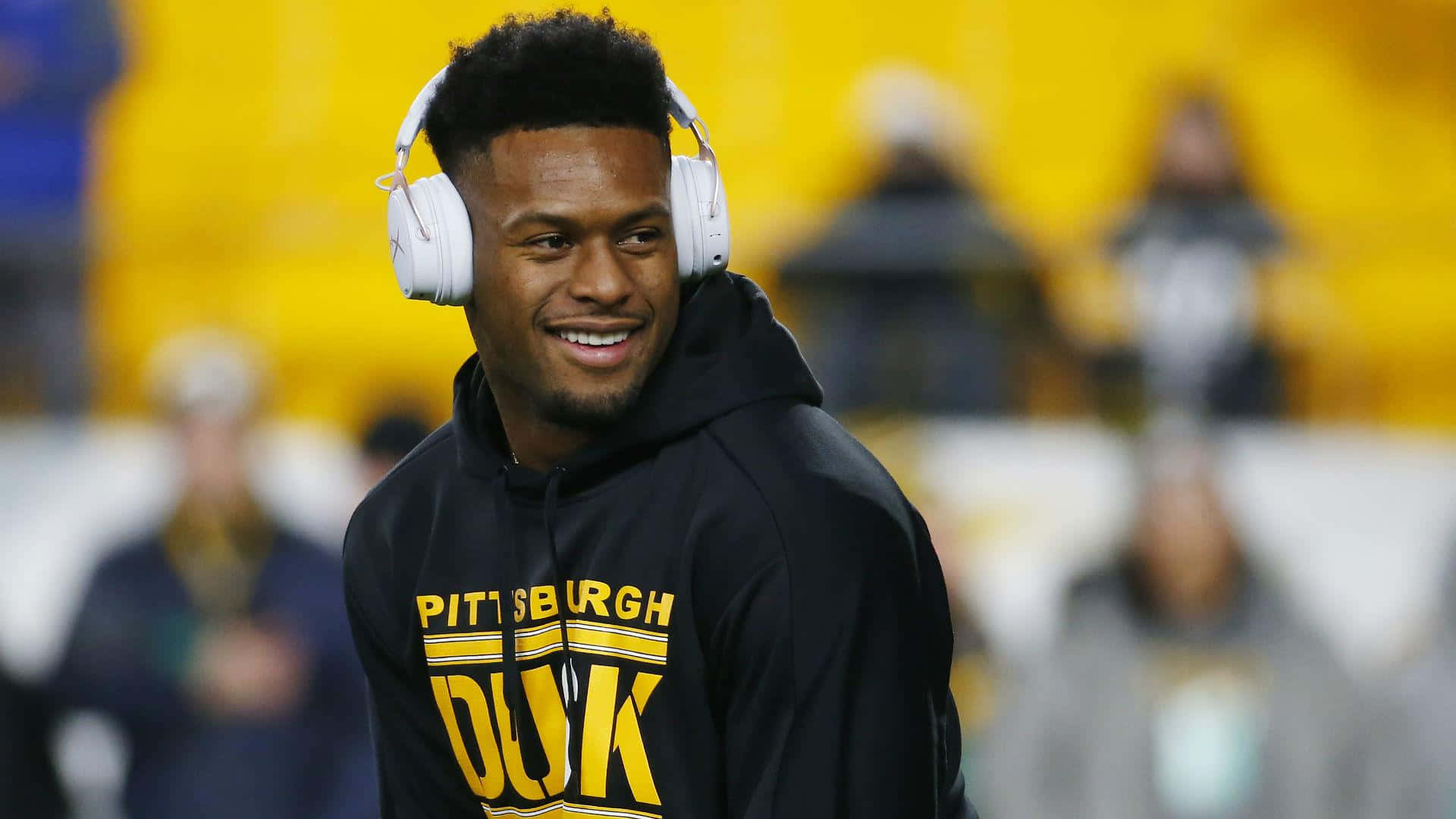 Juju Smith Schuster 桌布 壁紙 — 一位微笑的足球員穿著黑色連帽衫，上面有黃色 'PITTSBURGH DUK' 文字，戴著白色耳機，背景是模糊的體育場。