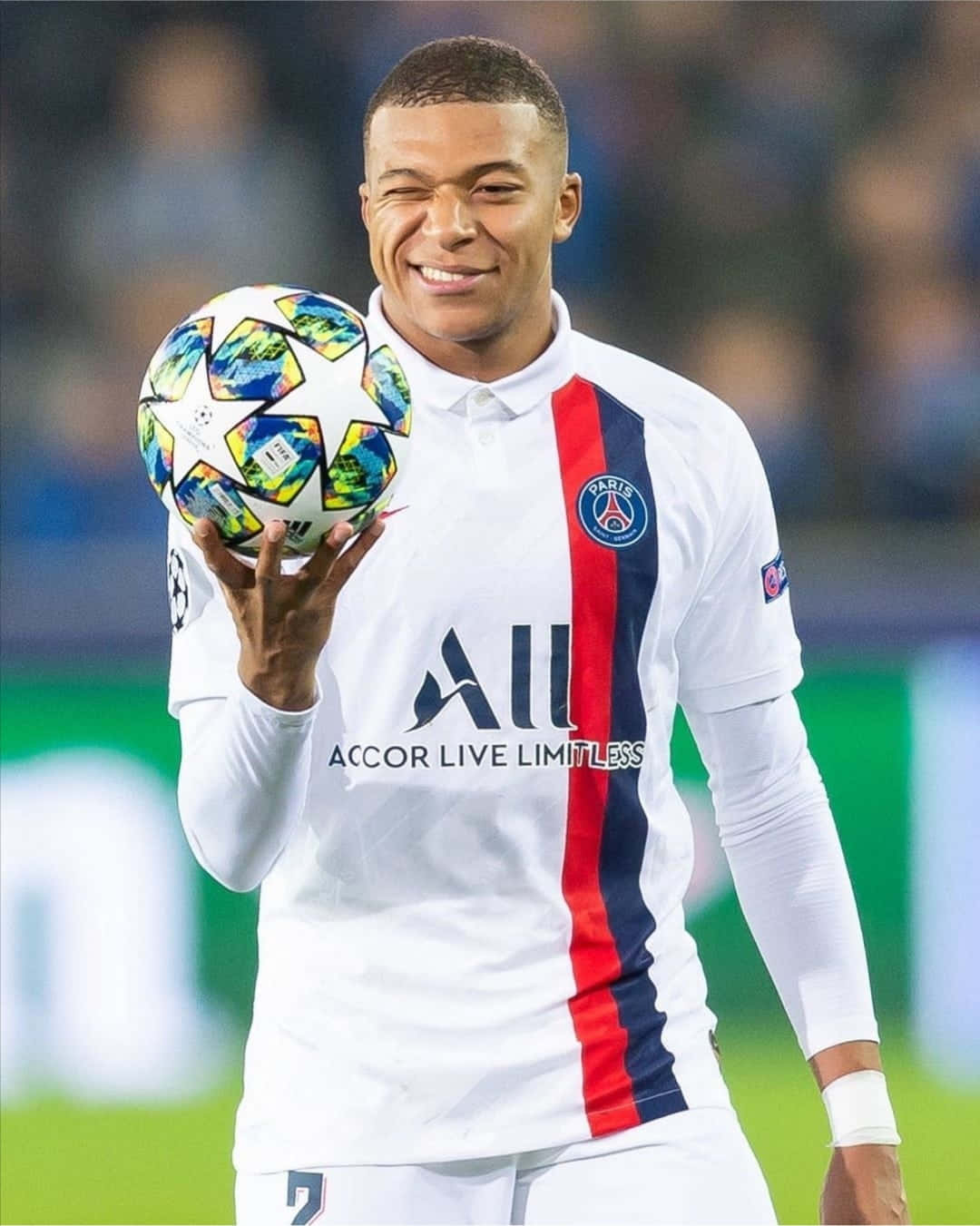 Mbappe 桌布 壁紙 — 一位微笑的足球員身穿巴黎聖日耳曼球衣，在球場上拿著色彩繽紛的歐冠足球。
